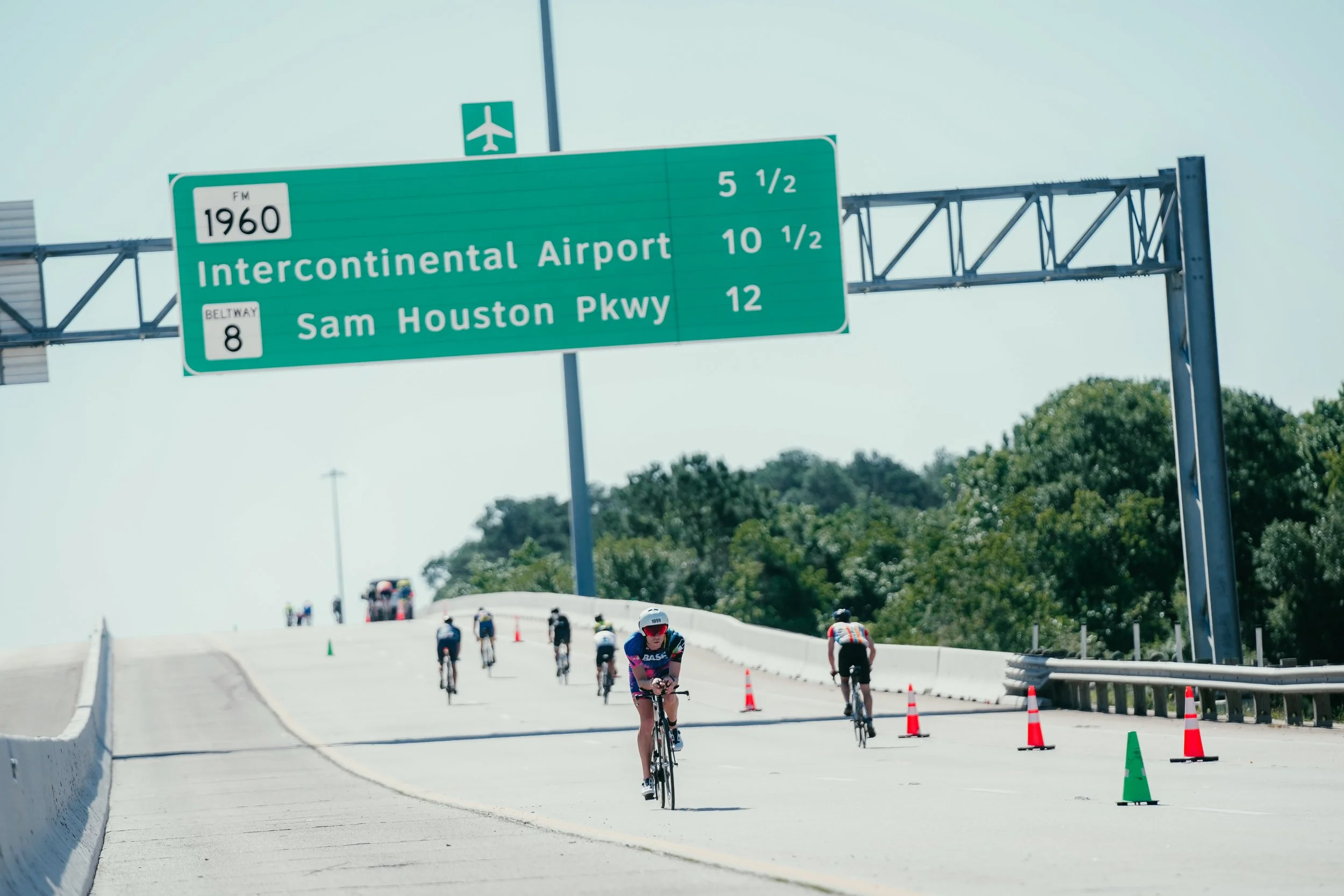 Cyclists participating in a race or event on a sunny day, riding on a road with orange and green cones, under a green highway sign that indicates directions to Intercontinental Airport and Sam Houston Parkway.