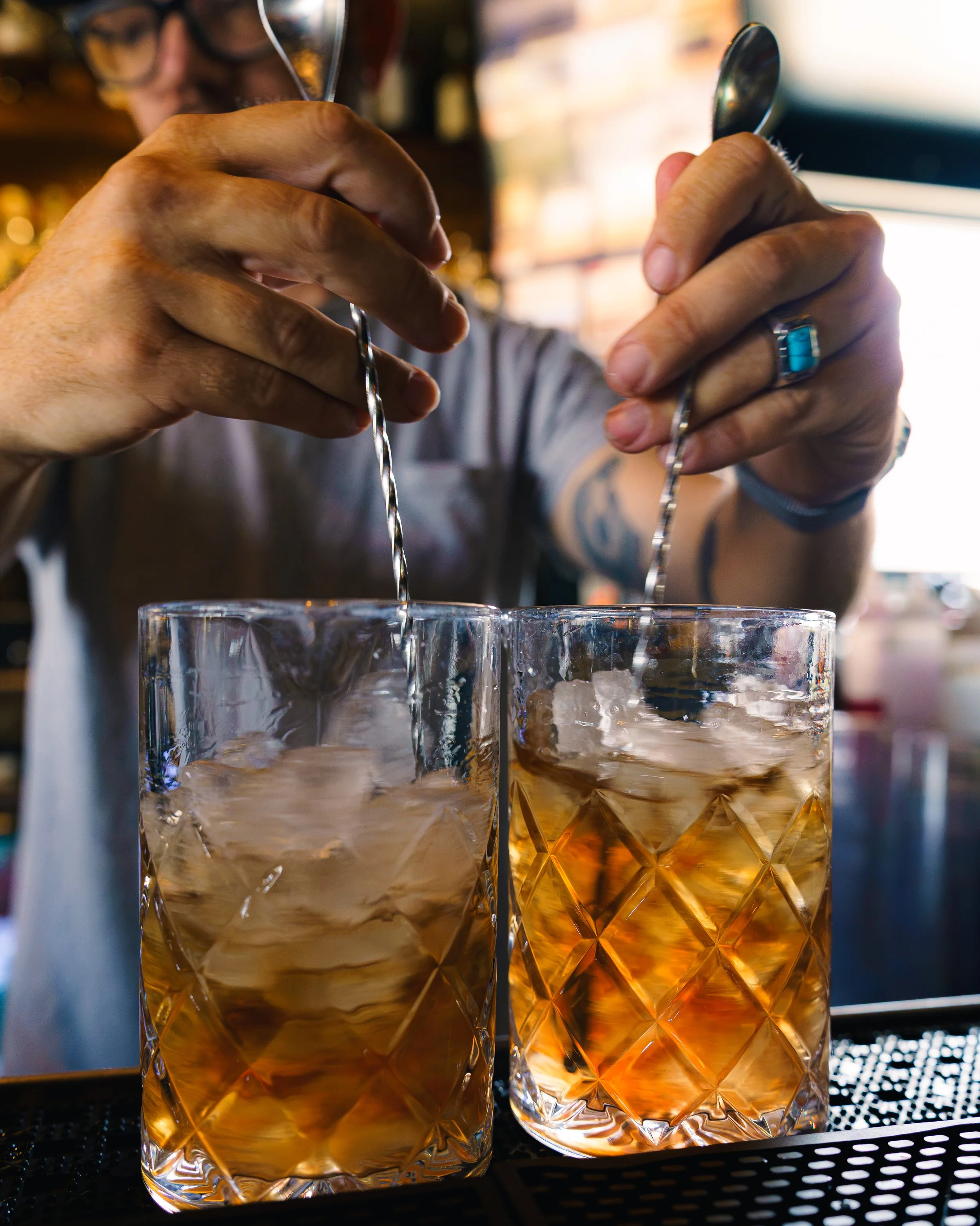 Two cocktails being stired in crystal glassware, set against a blurred background, with a person faintly visible in the distance.