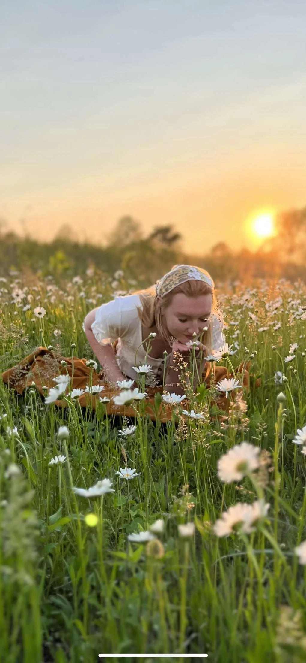 A woman with blonde hair, wearing a white shirt and a headscarf, is kneeling in a field of daisies during sunset, smelling the flowers.