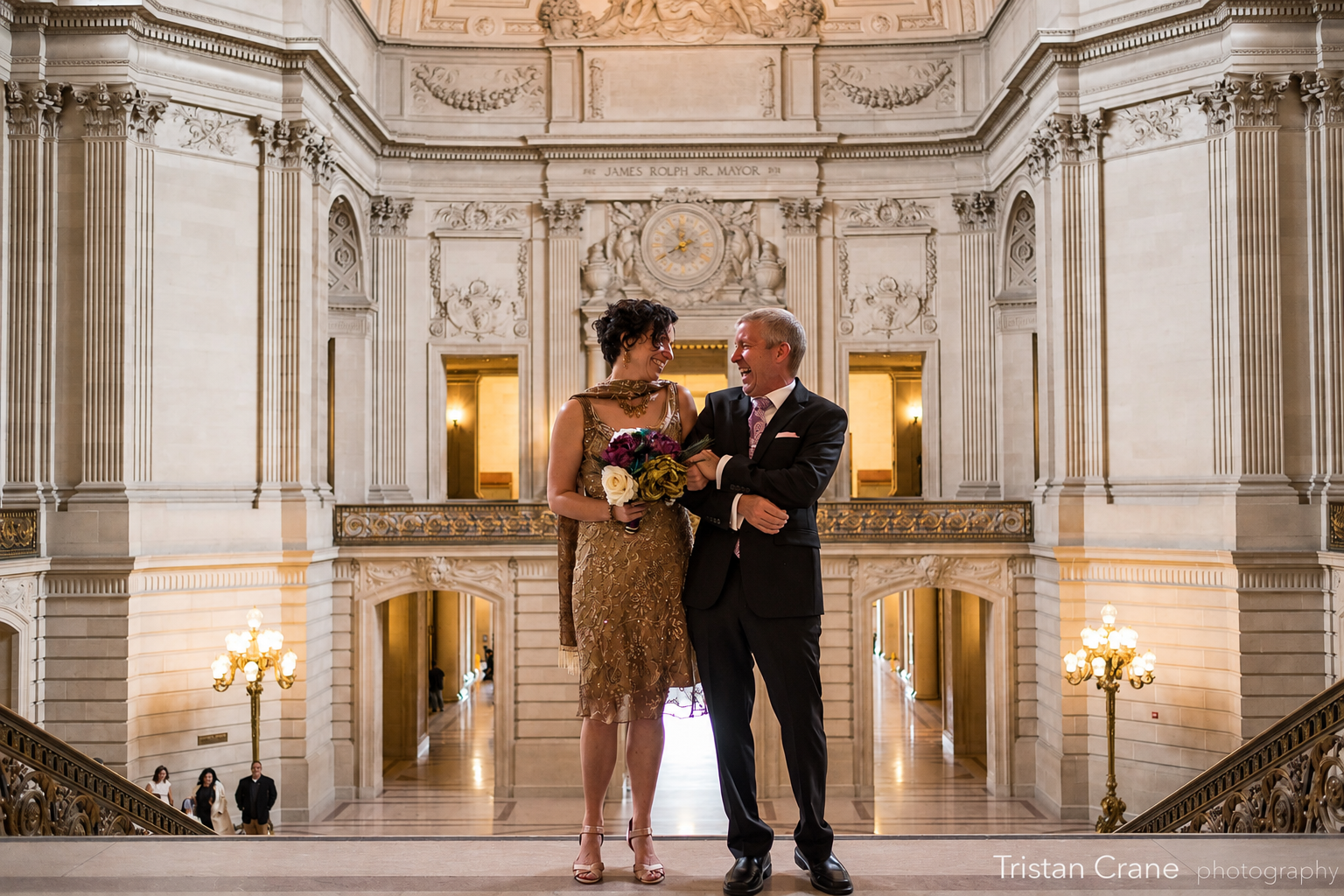 A man and woman dressed in formal attire standing inside a grand, ornate building. The woman is holding a bouquet of flowers and is wearing a gold dress, while the man is in a black suit and tie. They are smiling and looking at each other, standing on a staircase with elegant lighting and intricate architectural details behind them.