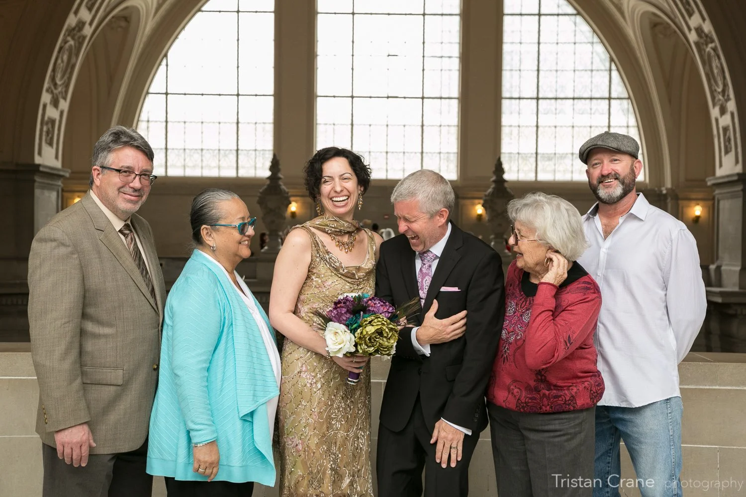 Group of seven diverse people smiling and laughing at a formal event in a grand hall, with large arched windows in the background. The woman in the center is holding a bouquet of flowers, and everyone is dressed in formal attire.