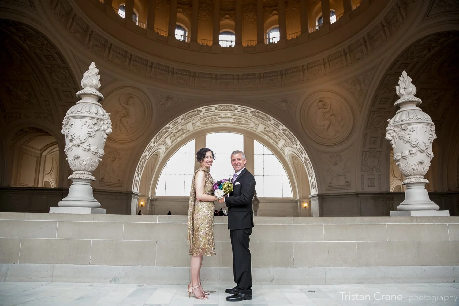 A couple dressed in formal attire stands in an ornate, grand hall with high ceilings, large arched windows, and decorative sculptures. The woman holds a bouquet of flowers, and both are smiling at the camera.