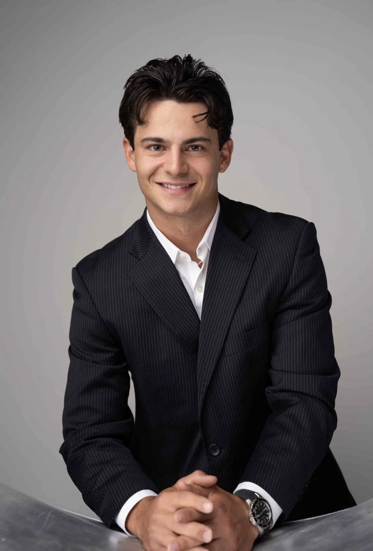A young man in a dark pinstripe suit with a white shirt, smiling and posing with hands clasped on a table, against a plain grey background.