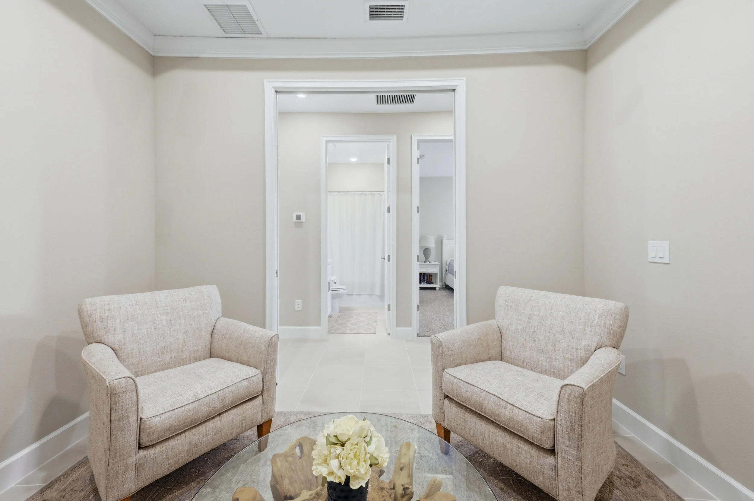 Living room with two beige armchairs facing each other, a glass coffee table with a flower arrangement in the center, and a hallway with two doorways leading to bedrooms in the background.