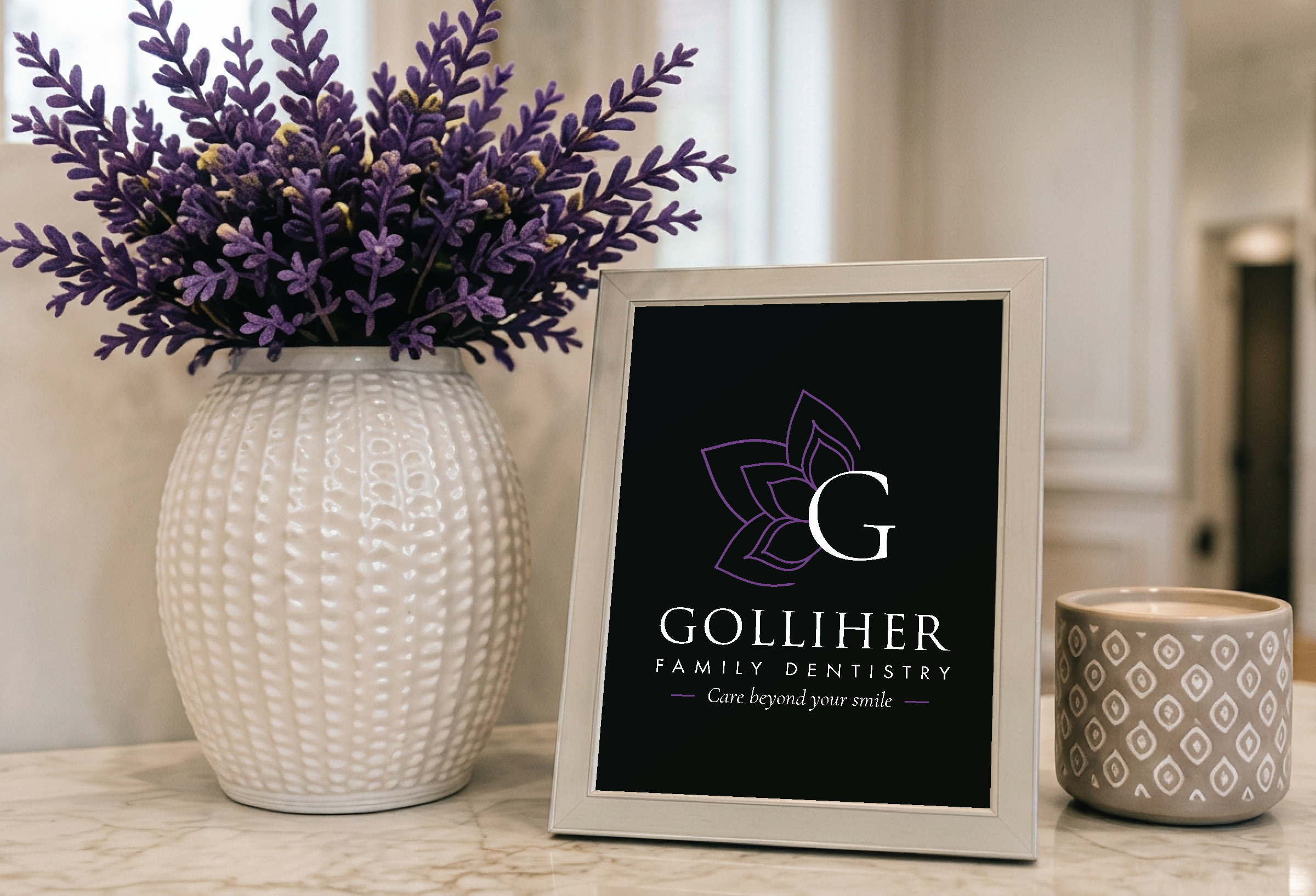 A white textured vase containing purple lavender flowers, a black name card for Gollihier Family Dentistry with a white and purple logo, and a patterned beige candle, all arranged on a marble surface in a bright space.