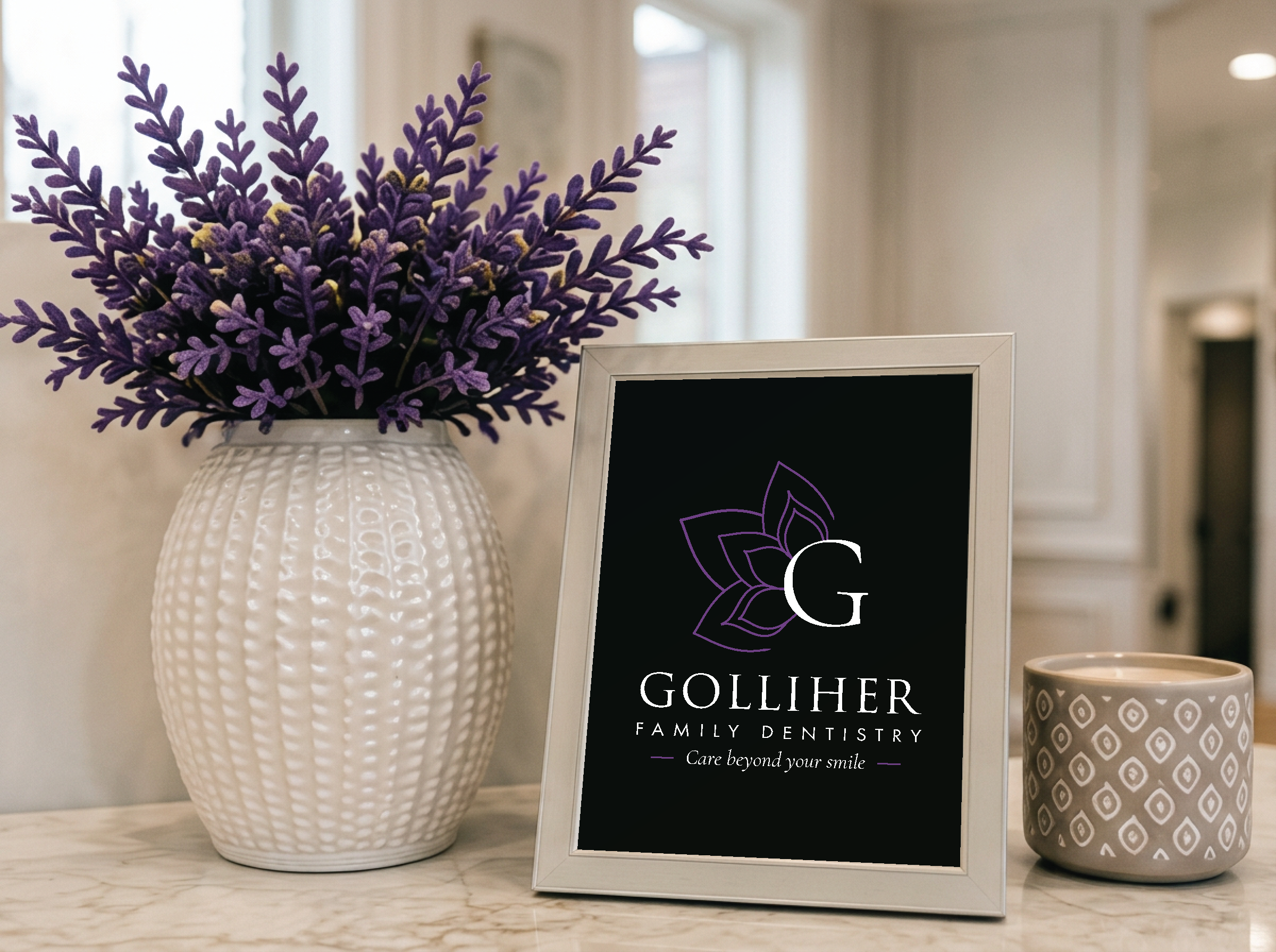 A white textured vase with purple lavender flowers next to a black framed sign for Gollihner Family Dentistry, on a marble countertop. The sign includes a large decorative flower, the letter G, and the tagline 'Care beyond your smile.' A small patterned ceramic cup is partially visible on the right.