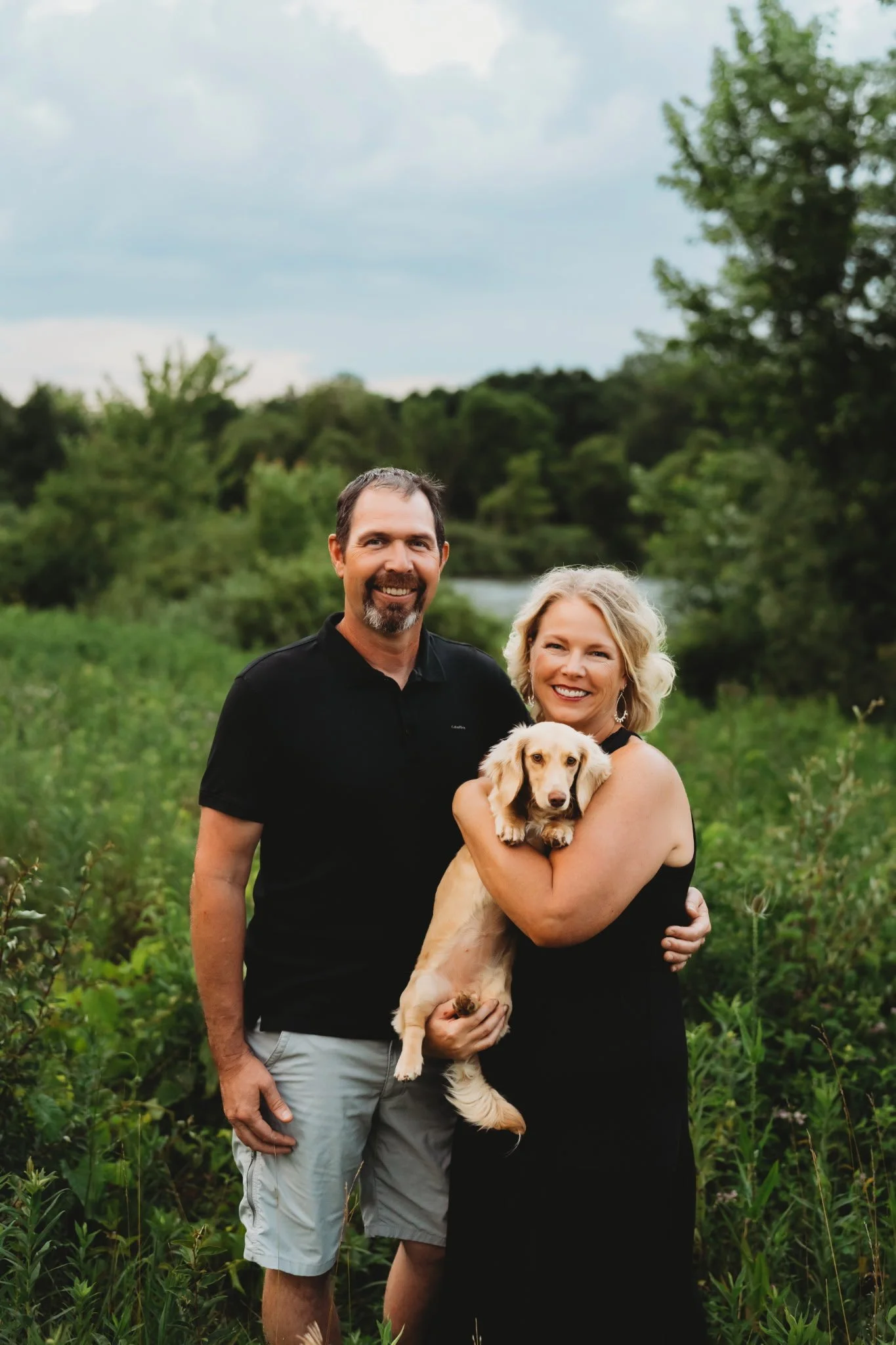 A smiling couple with a dog stands outdoors in a green field with trees and a cloudy sky in the background.