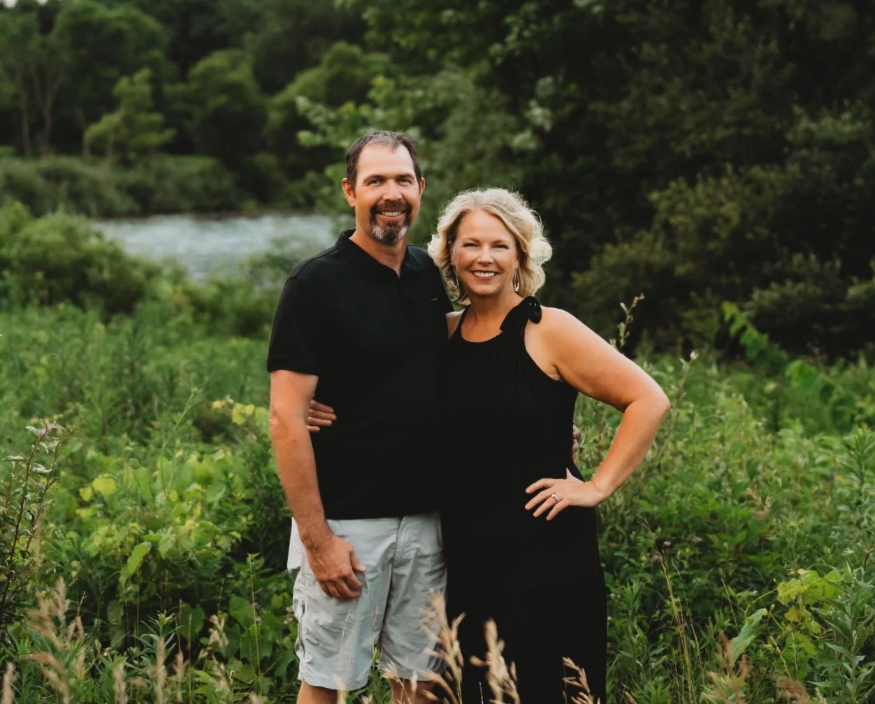 A smiling couple standing outdoors in a lush green area with a river in the background. The man has short dark hair and a beard, wearing a black polo shirt and light-colored shorts. The woman has blonde curly hair, wearing a black sleeveless dress with bow details on the shoulders. They are posing together with arms around each other.