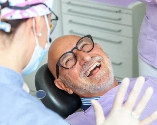 A man with glasses lying back in a dental chair, smiling and laughing, while a dental worker attends to him.