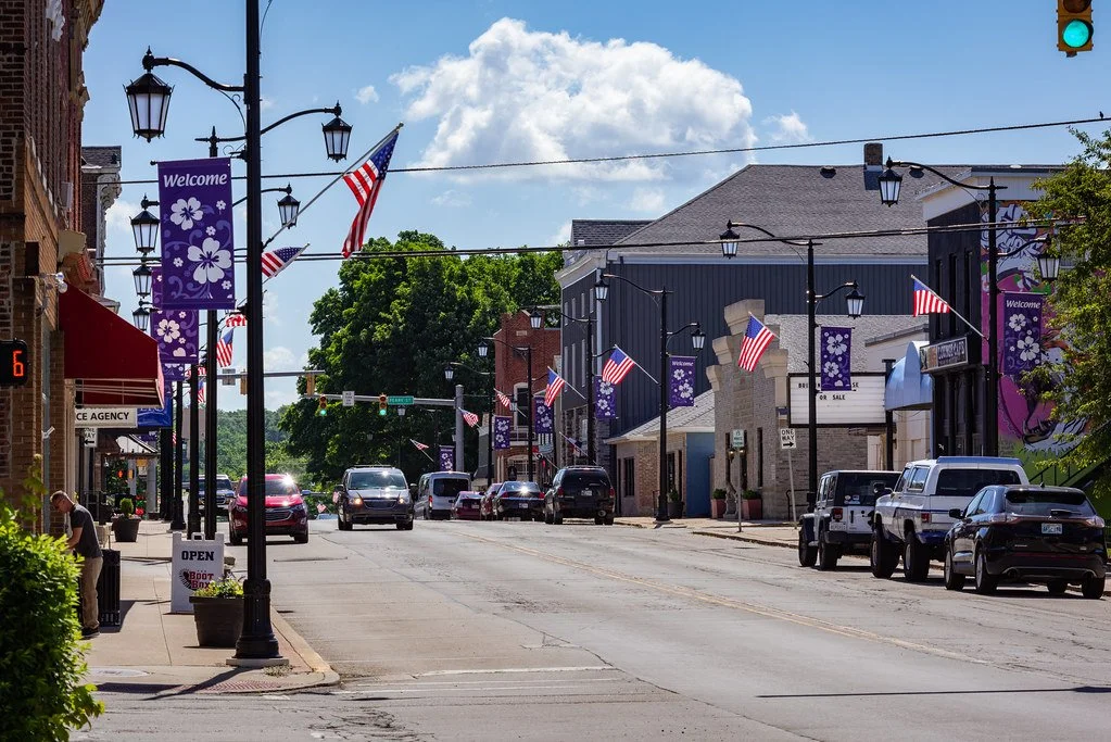 Downtown main street with American flags and purple welcome banners, cars parked along the side, and a partly cloudy sky.