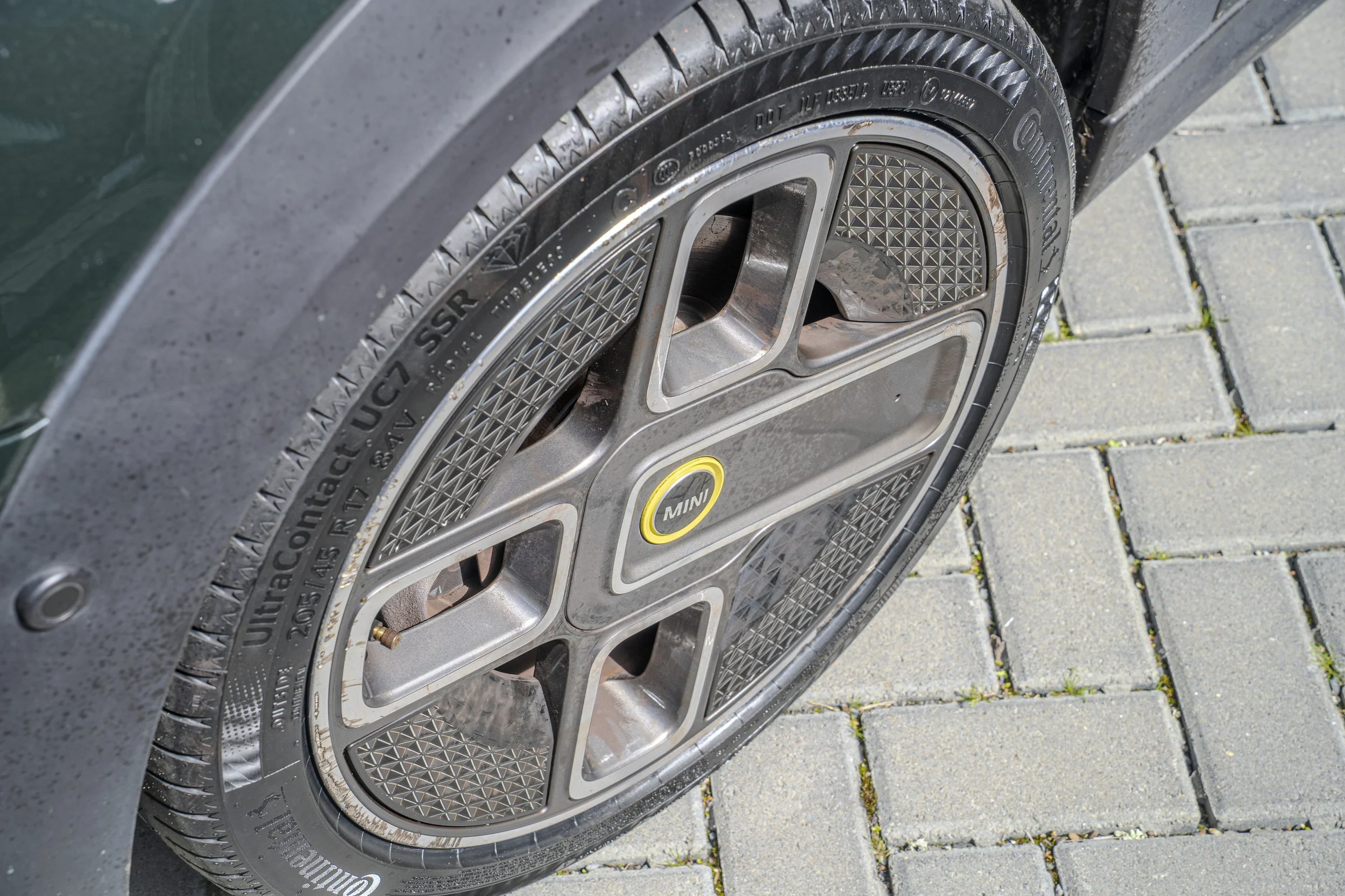 Close-up of a car's wheel with a Continental tire and a Mini logo on the wheel hub, parked on a paved surface with gray bricks.