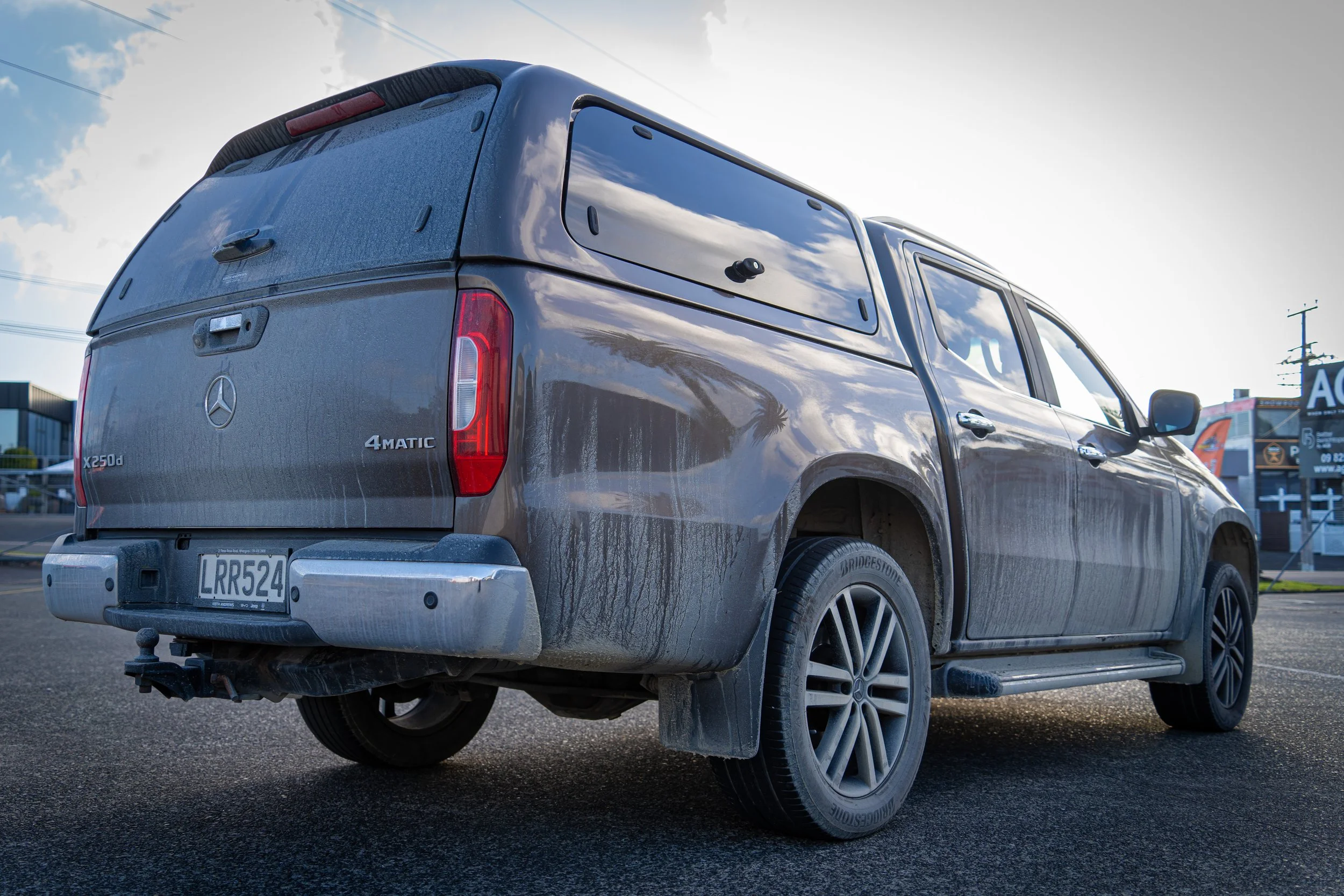 A dark gray Mercedes-Benz X250d 4MATIC pickup truck parked outdoors during daytime with a cloudy sky, power lines, and signage visible in the background.