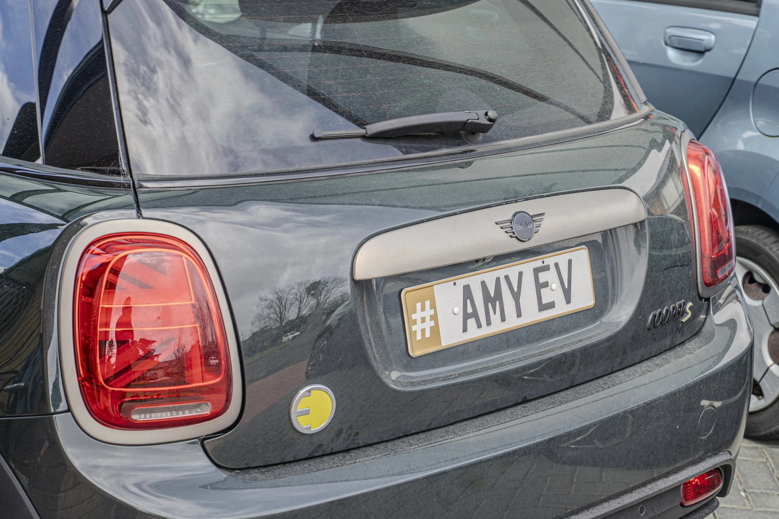 The rear end of a black Mini Cooper S car parked in a parking lot, showing its tail lights, rear window, and a license plate with the hashtag #AMY-EV.
