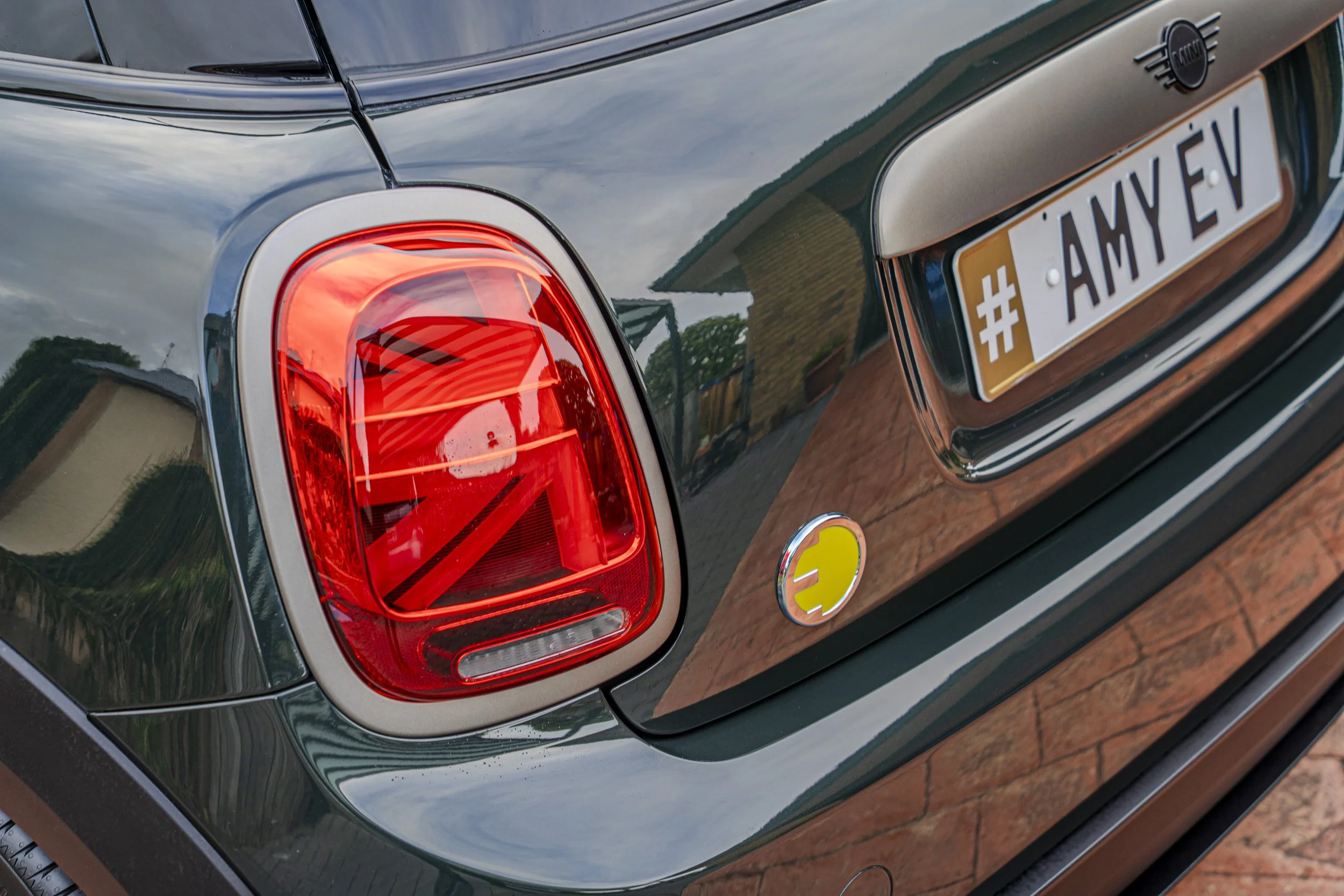 Close-up of the rear end of a black Mini Cooper car featuring a red tail light, a brown wooden panel, a custom license plate with the text '#AMY EV', and a small round badge with a yellow smiley face.