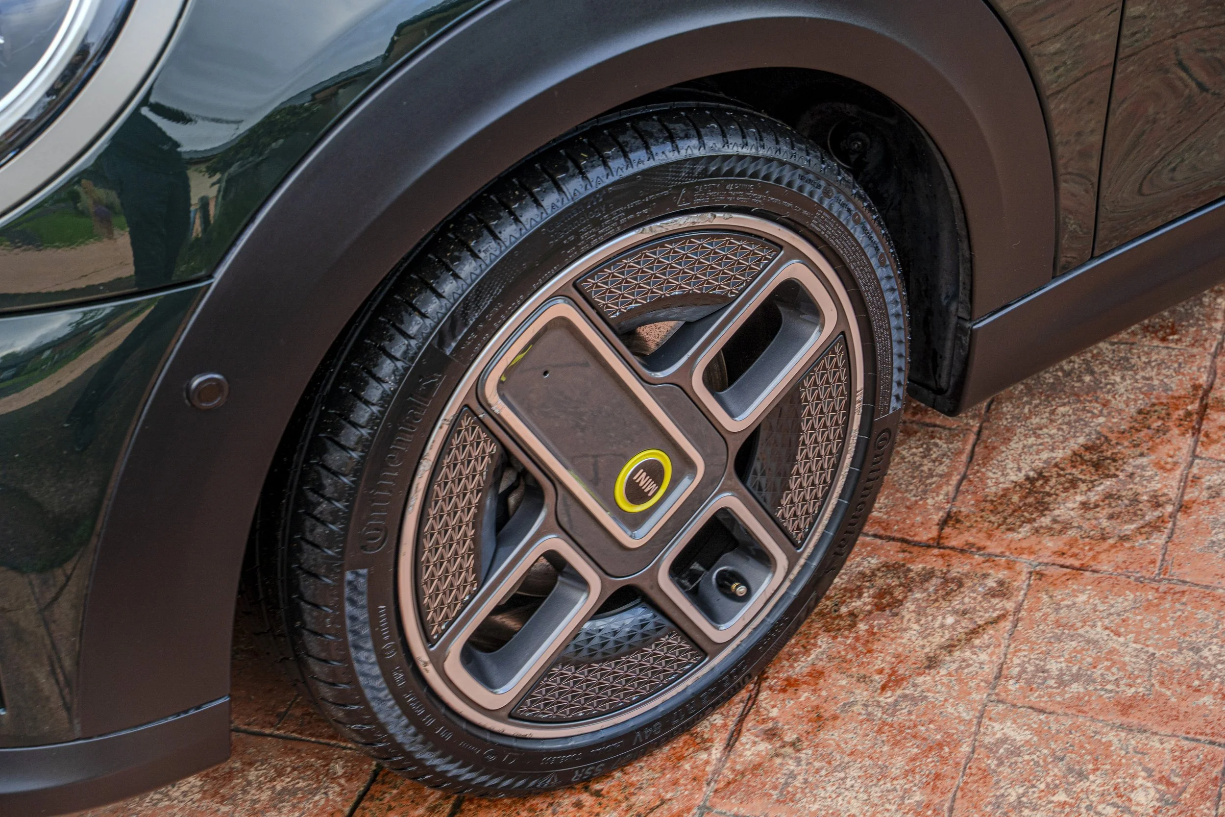 Close-up of a car's front wheel, showing the tire, rim, and part of the car's body, parked on a brick driveway.