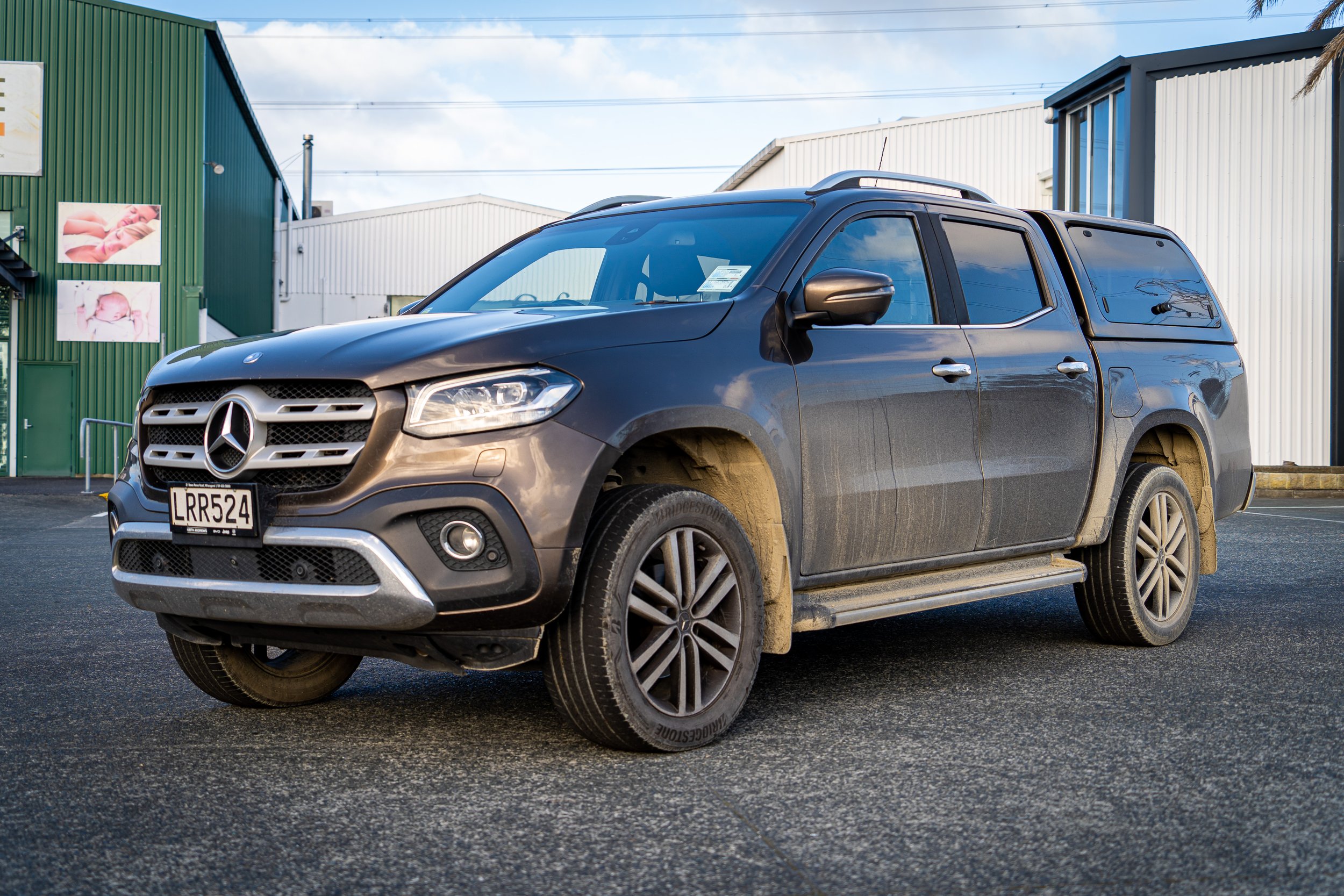 A metallic gray Mercedes-Benz SUV parked outside in front of industrial buildings with metal siding. The vehicle has visible mud on the tires and lower body, and the license plate reads LRR524. The background features blue sky with some clouds and po