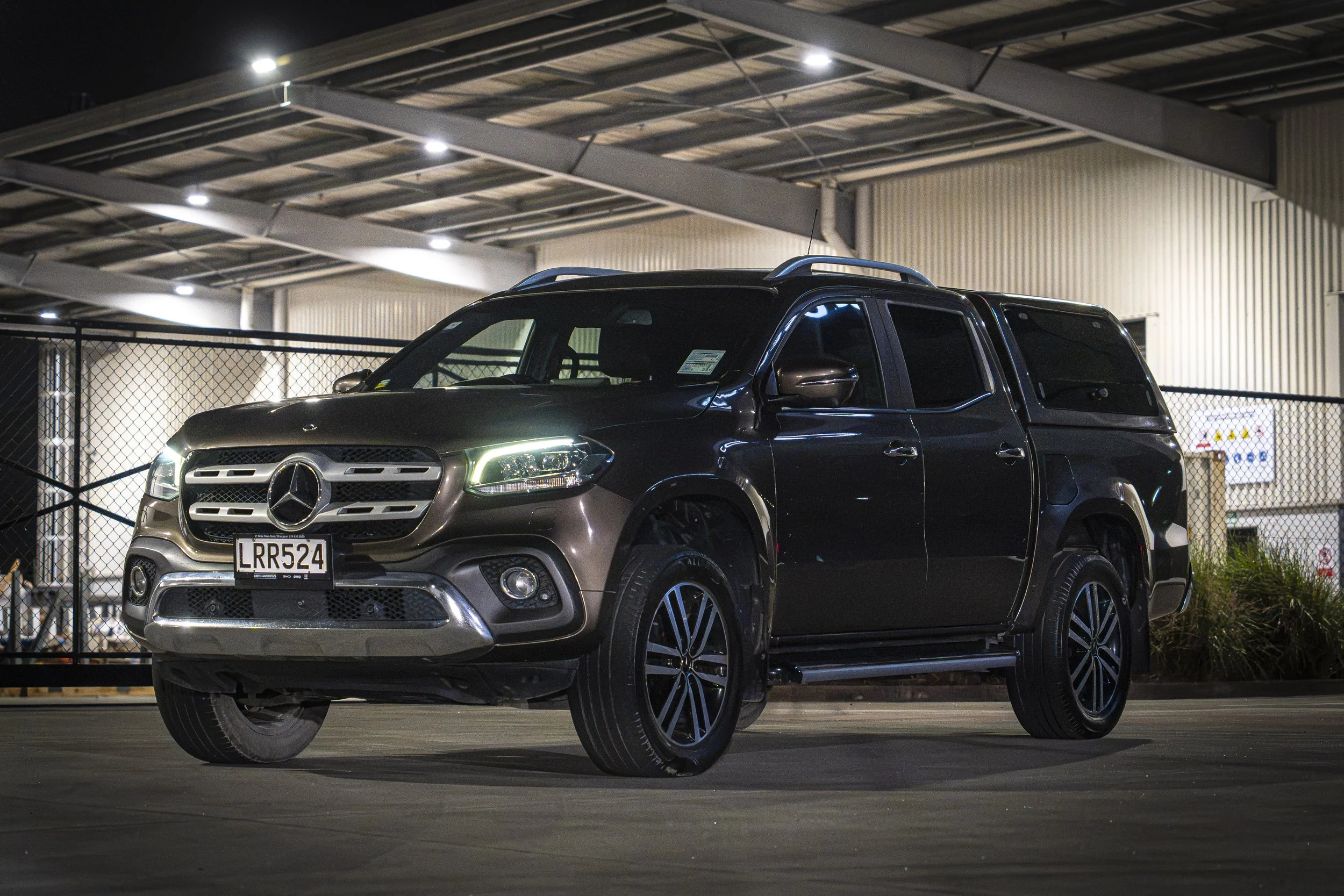 A black Mercedes-Benz pickup truck parked in a well-lit outdoor parking lot at night, with a building and a chain-link fence in the background.