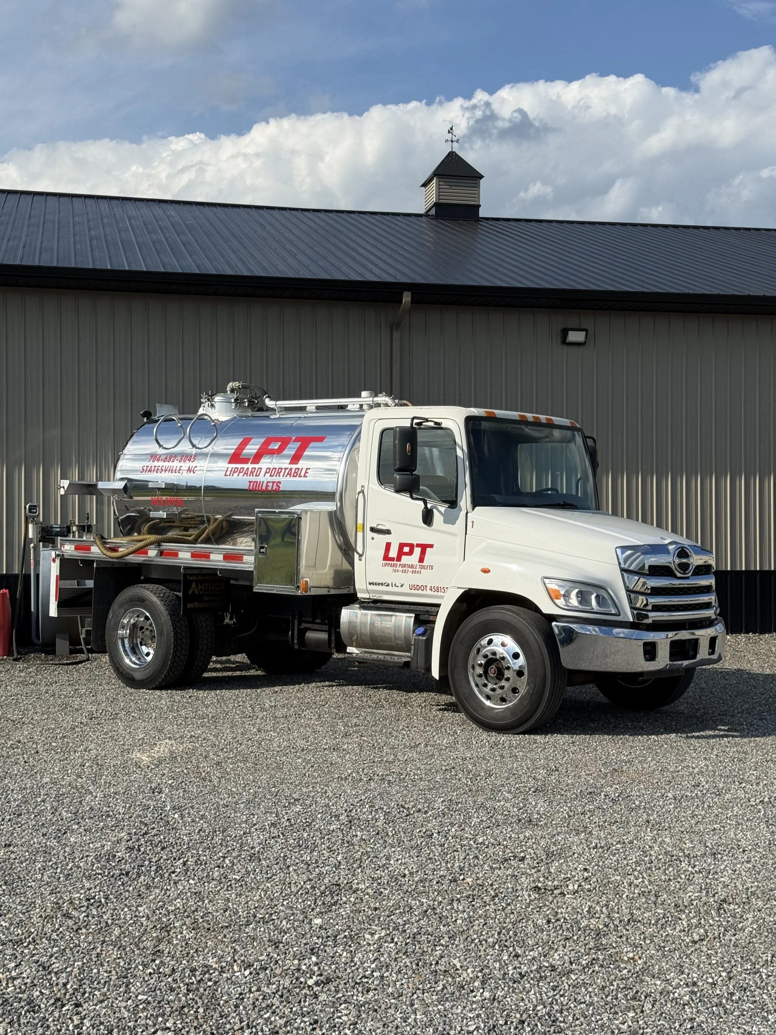 A white sanitation truck with red lettering parked outside a gray building with a metal roof and small cupola, under a partly cloudy sky.