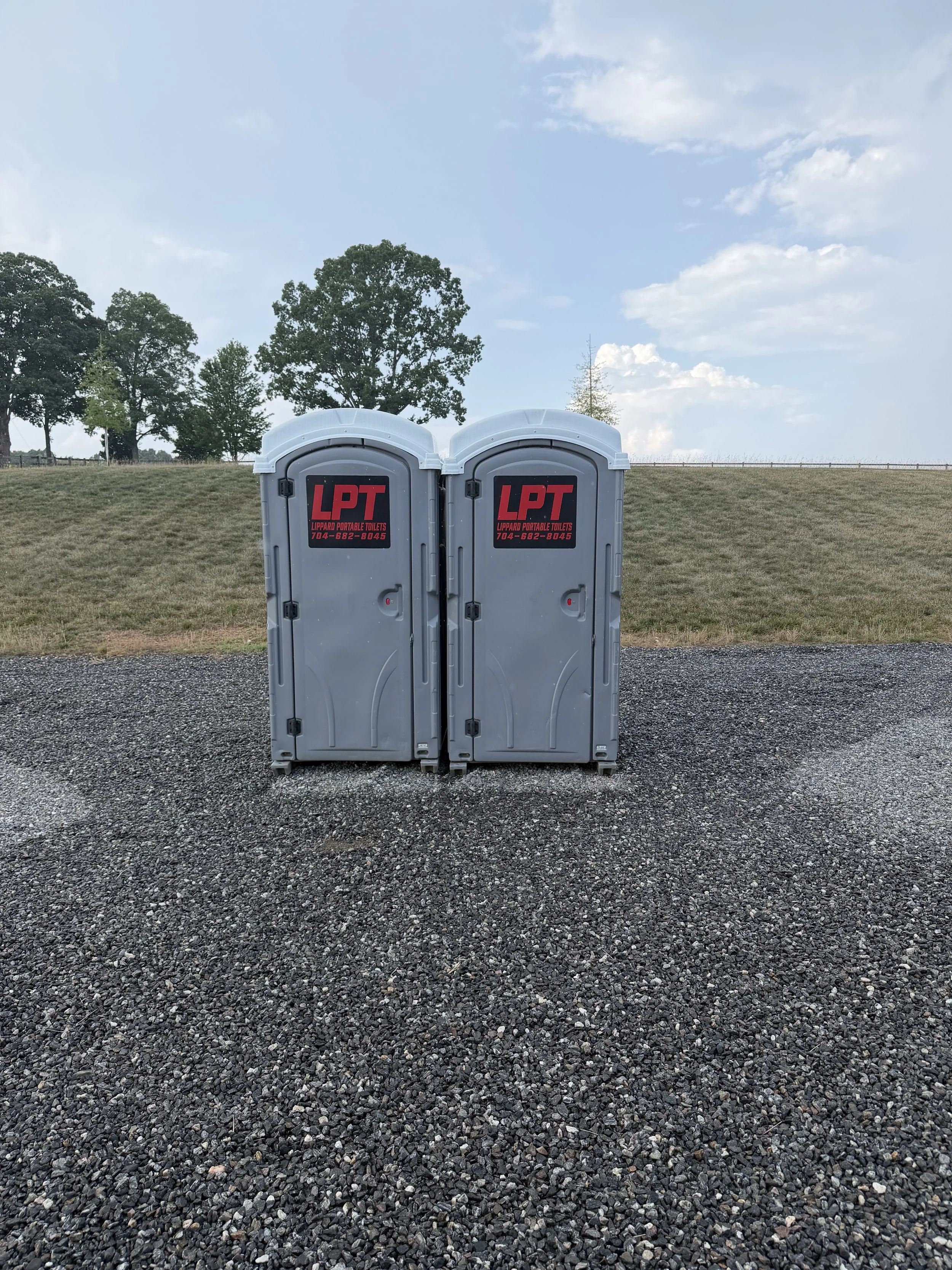 Two portable toilets with black and red labels standing on a gravel ground in front of a grassy field with trees under a blue sky.