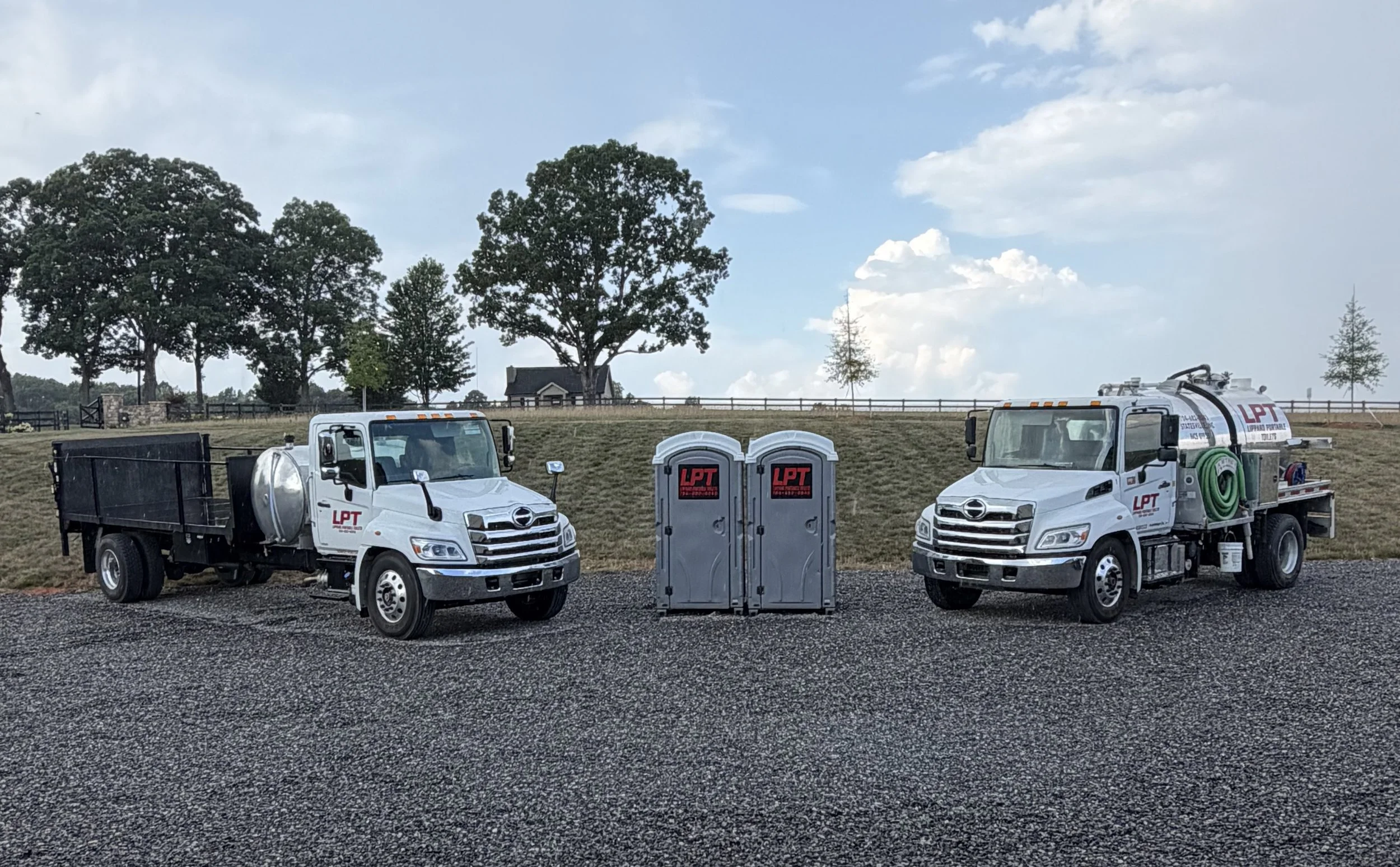 Two white utility trucks with company logos are parked in front of portable toilets and trees in a rural area.