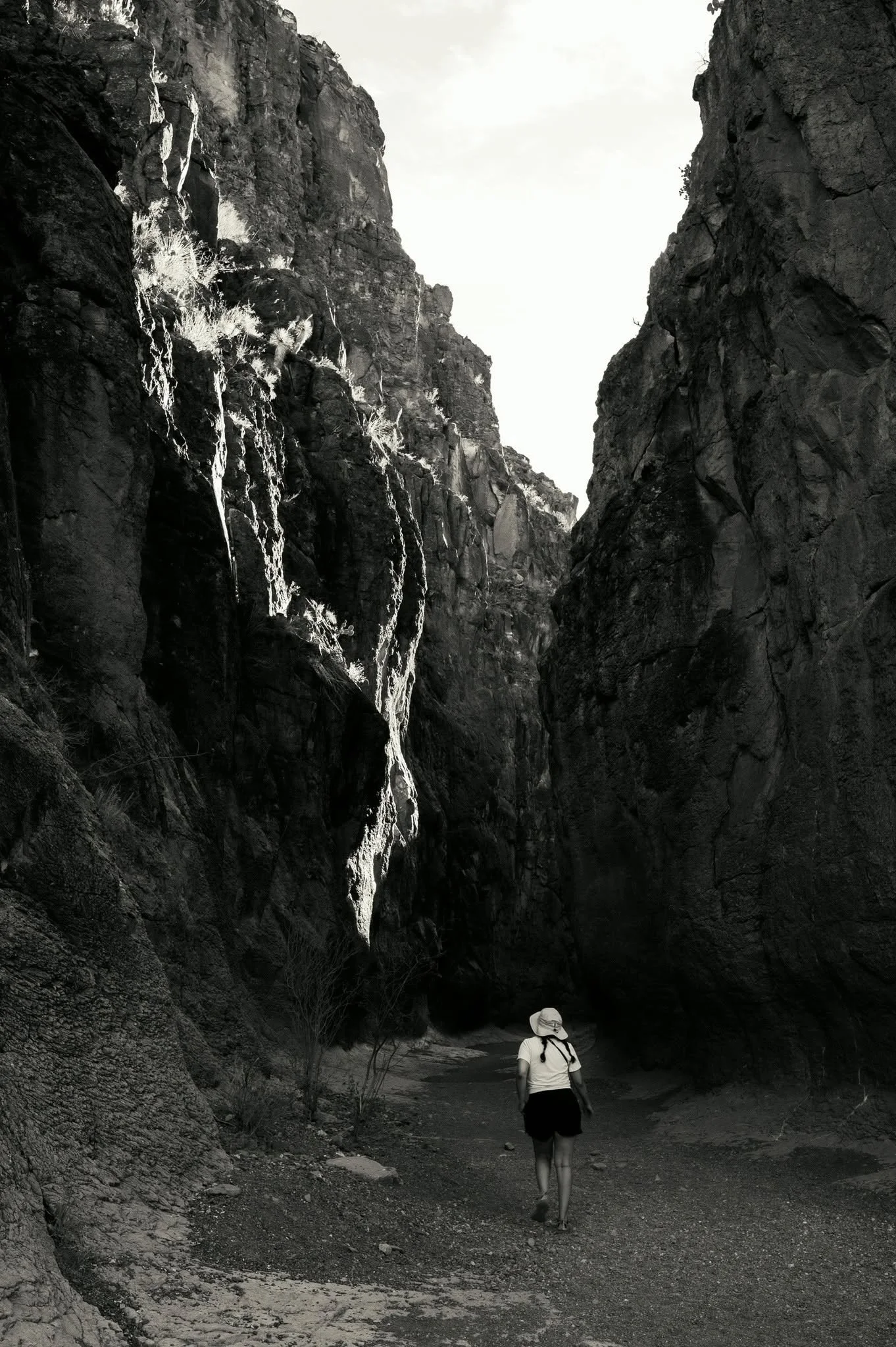 A person walking through a narrow canyon with high rocky walls on both sides. The person is wearing a hat and backpack and appears to be exploring or hiking. The canyon walls are steep and rugged with sparse vegetation.
