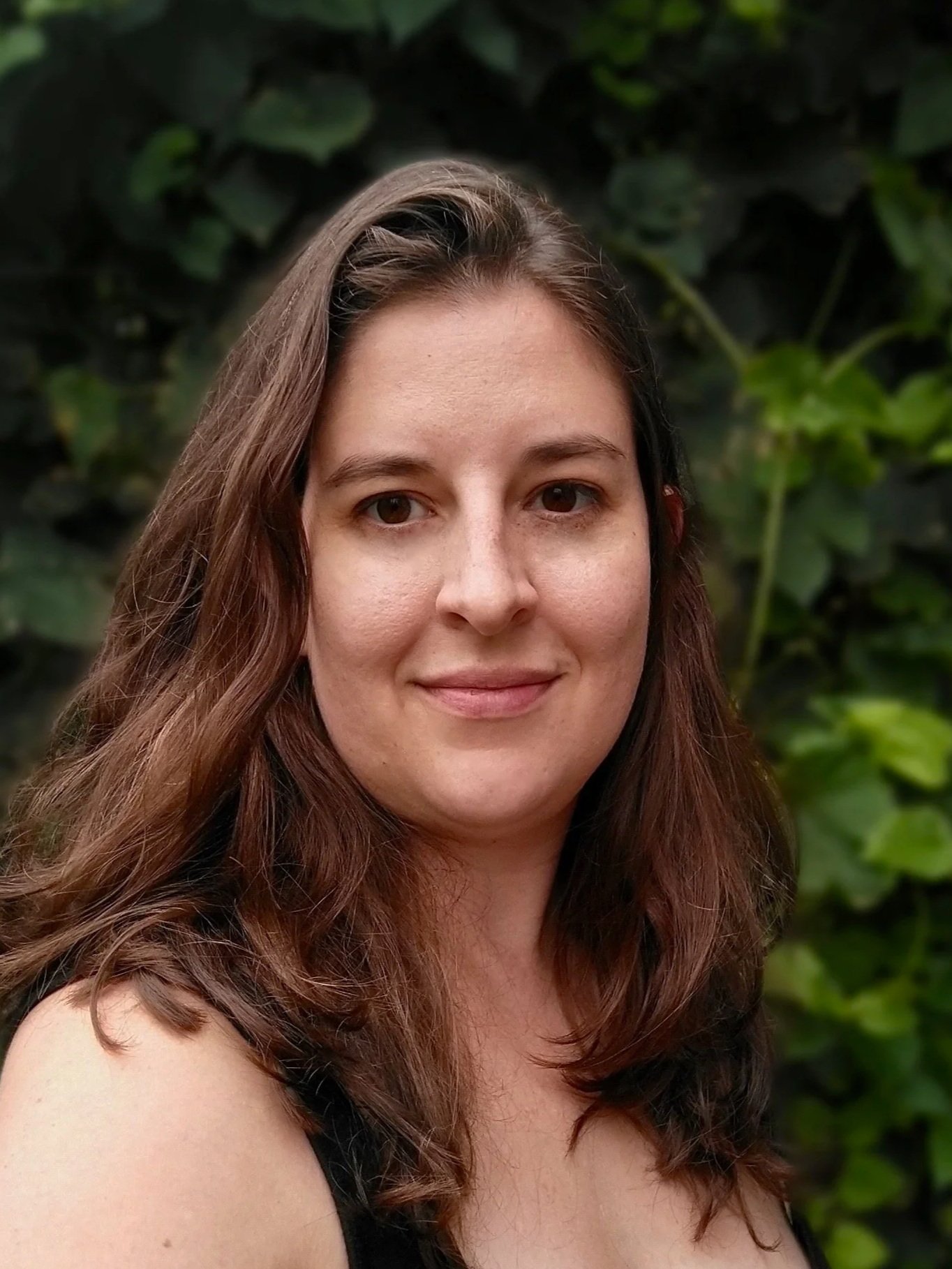 Close-up portrait of a woman with long, wavy brown hair, smiling slightly in front of a blurred background of green foliage.
