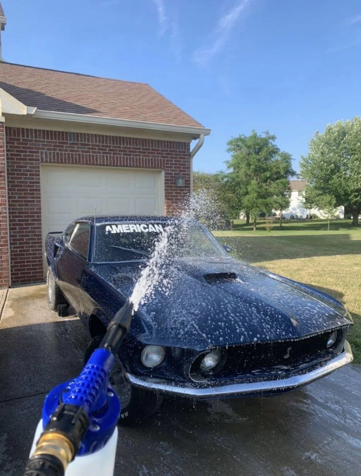Black Chevrolet Corvette car being washed in the driveway of a suburban home on a bright, sunny day with a blue sky and green trees in the background.