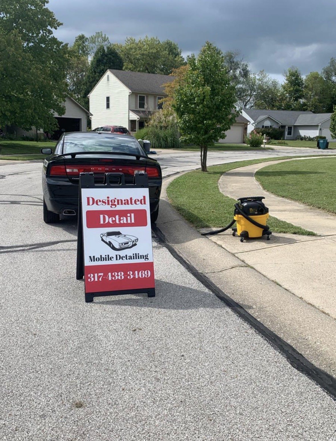 A black Dodge car parked on a neighborhood street next to a sidewalk. There is a sign in front of the car that reads "Designated Detail" and "Mobile Detailing" with a phone number. A yellow vacuum cleaner is on the sidewalk nearby. Residential houses and trees are visible in the background under a cloudy sky.