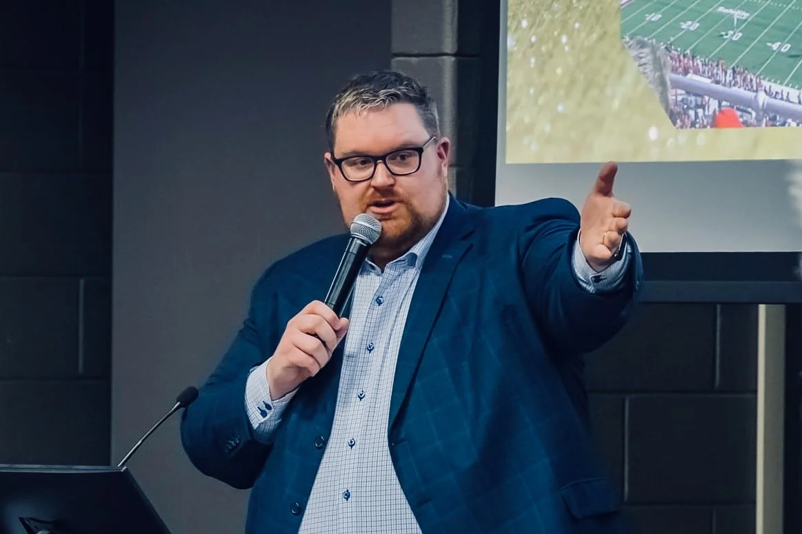 A man in a blue suit and glasses holding a microphone and gesturing with his right hand during a presentation