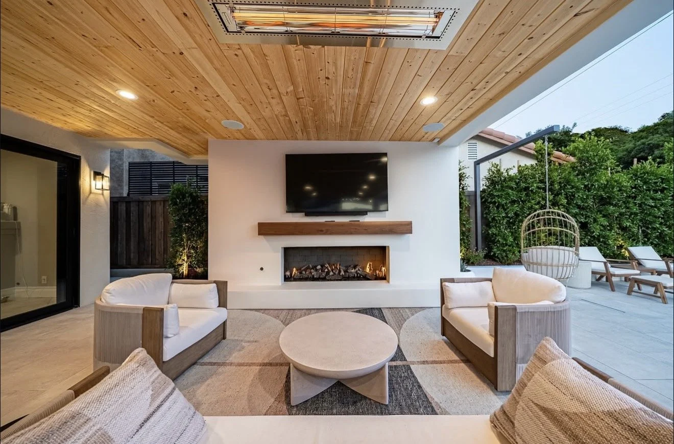 Outdoor patio area with seating, a fireplace, a wall-mounted TV, a wood-paneled ceiling, and a hanging egg chair surrounded by greenery.