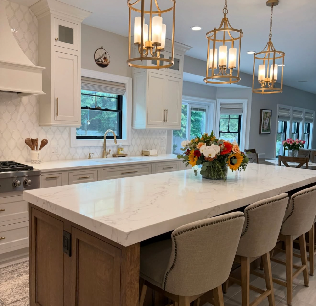 Modern kitchen with white marble island, white cabinets, large window, and floral centerpieces hanging from gold chandeliers.