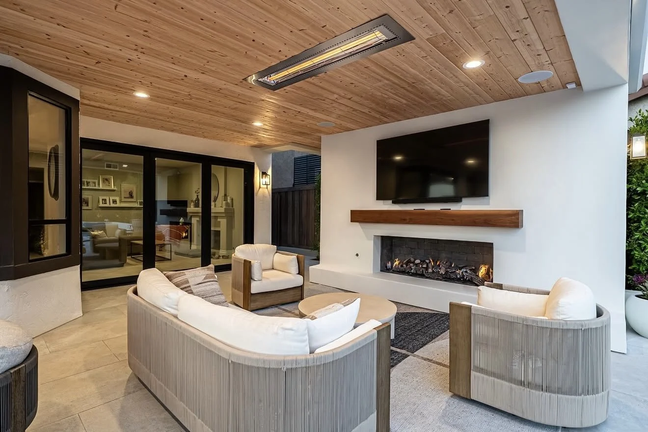 Living room with curved beige sofas, a wooden coffee table, a wall-mounted TV, and a fireplace below the TV. The ceiling is made of wood planks with recessed lighting, and there are large sliding glass doors leading to another room.