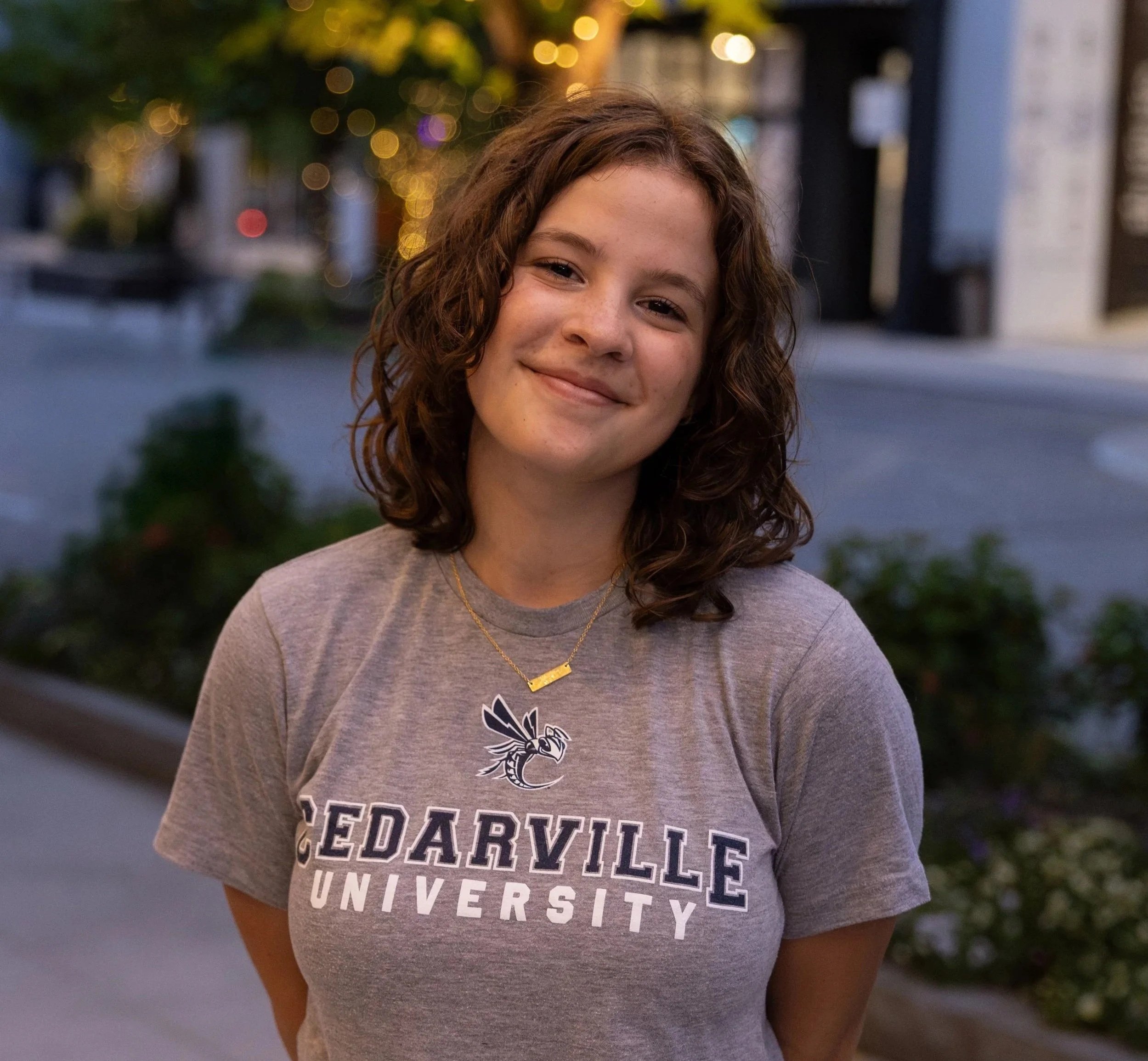 Close-up of a smiling girl with curly hair wearing a gray t-shirt with university names and a gold necklace, outdoors at dusk with a background of blurred lights and trees.