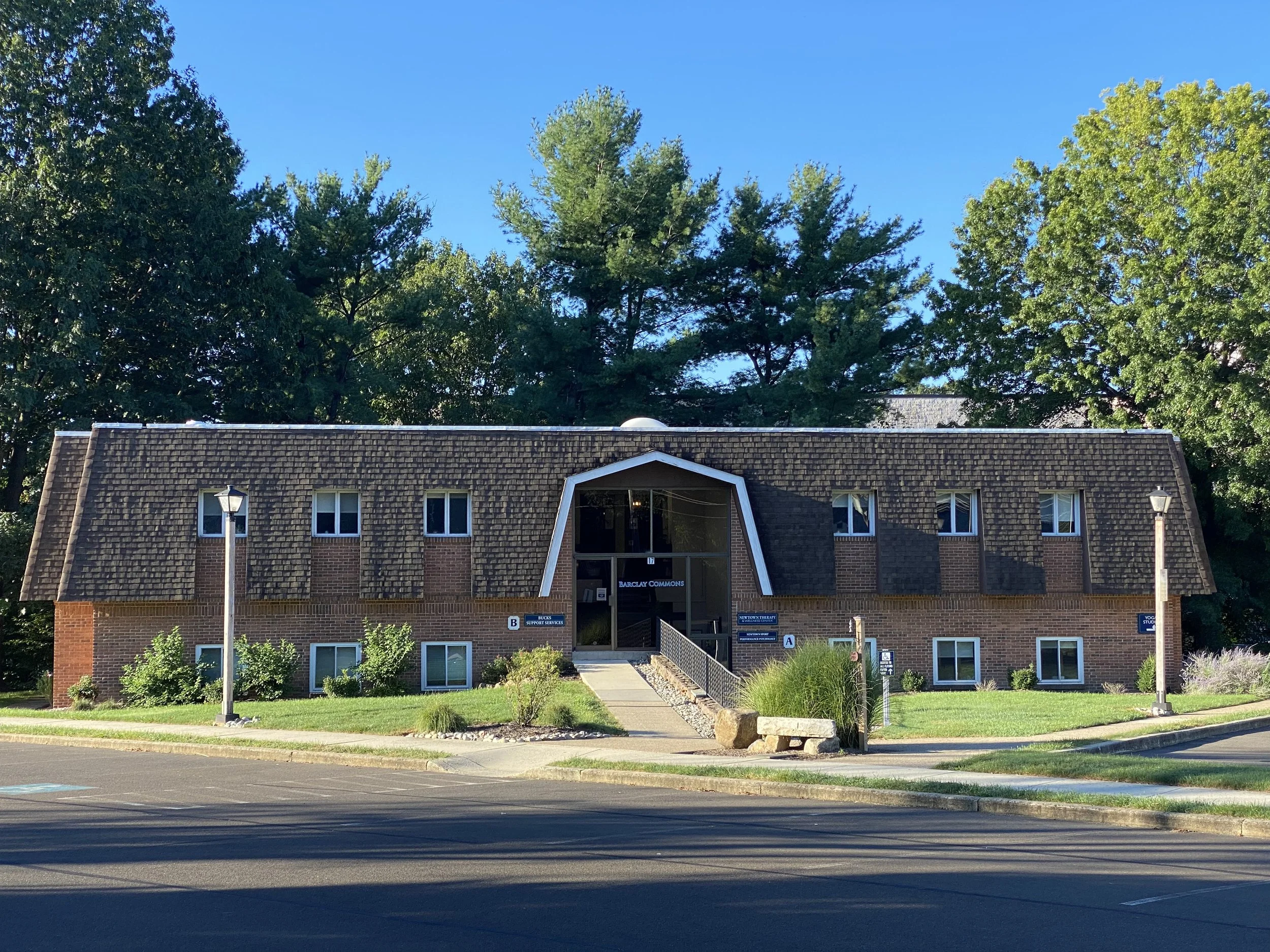 A two-story brick building with a sloped roof, multiple windows, and signage indicating it is the Barclay Commons support services building. The building is surrounded by a green lawn, bushes, and trees. A sidewalk leads up to the entrance, with two lampposts on either side of the walkway. The sky is clear and blue.
