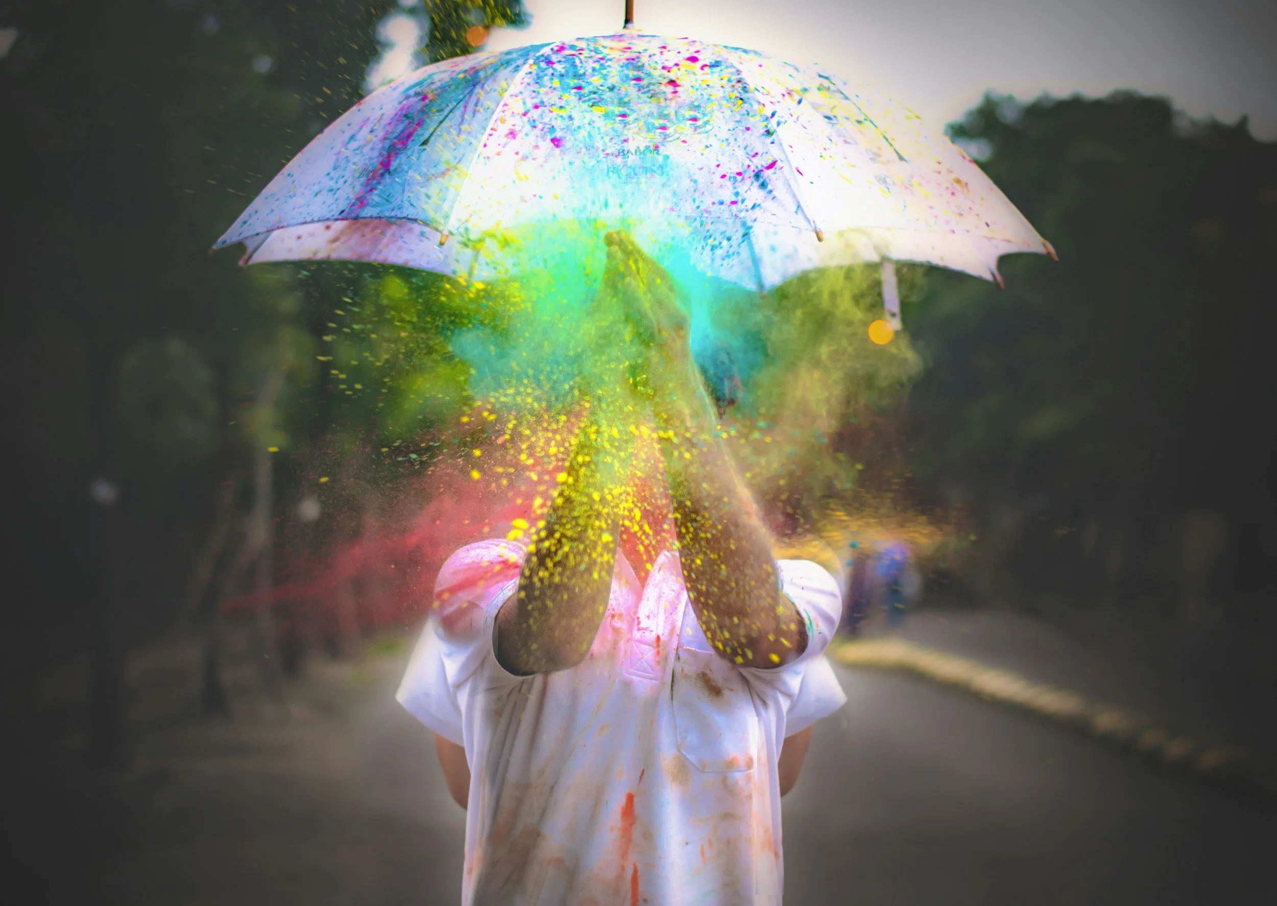 Child holding a colorful umbrella that is splattered with bright paint, standing outdoors with trees in the background.