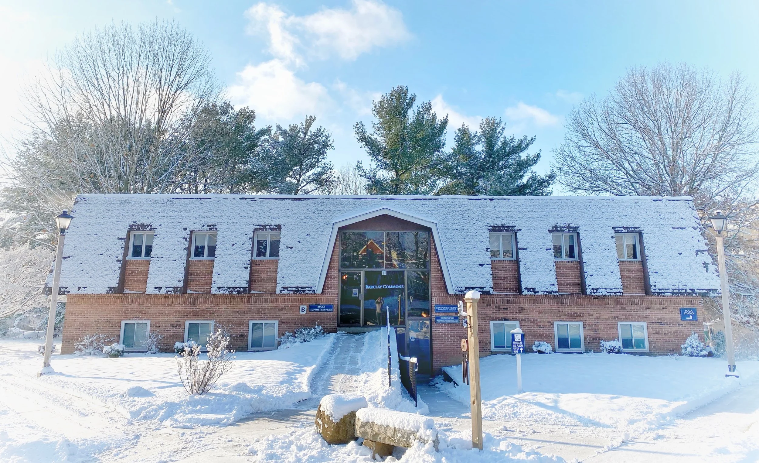 A brick building with snow-covered roof and windows, with trees in the background and a blue sky. Signage indicates it is Barclays Commons and a yoga studio, with accessibility and support services signs outside.