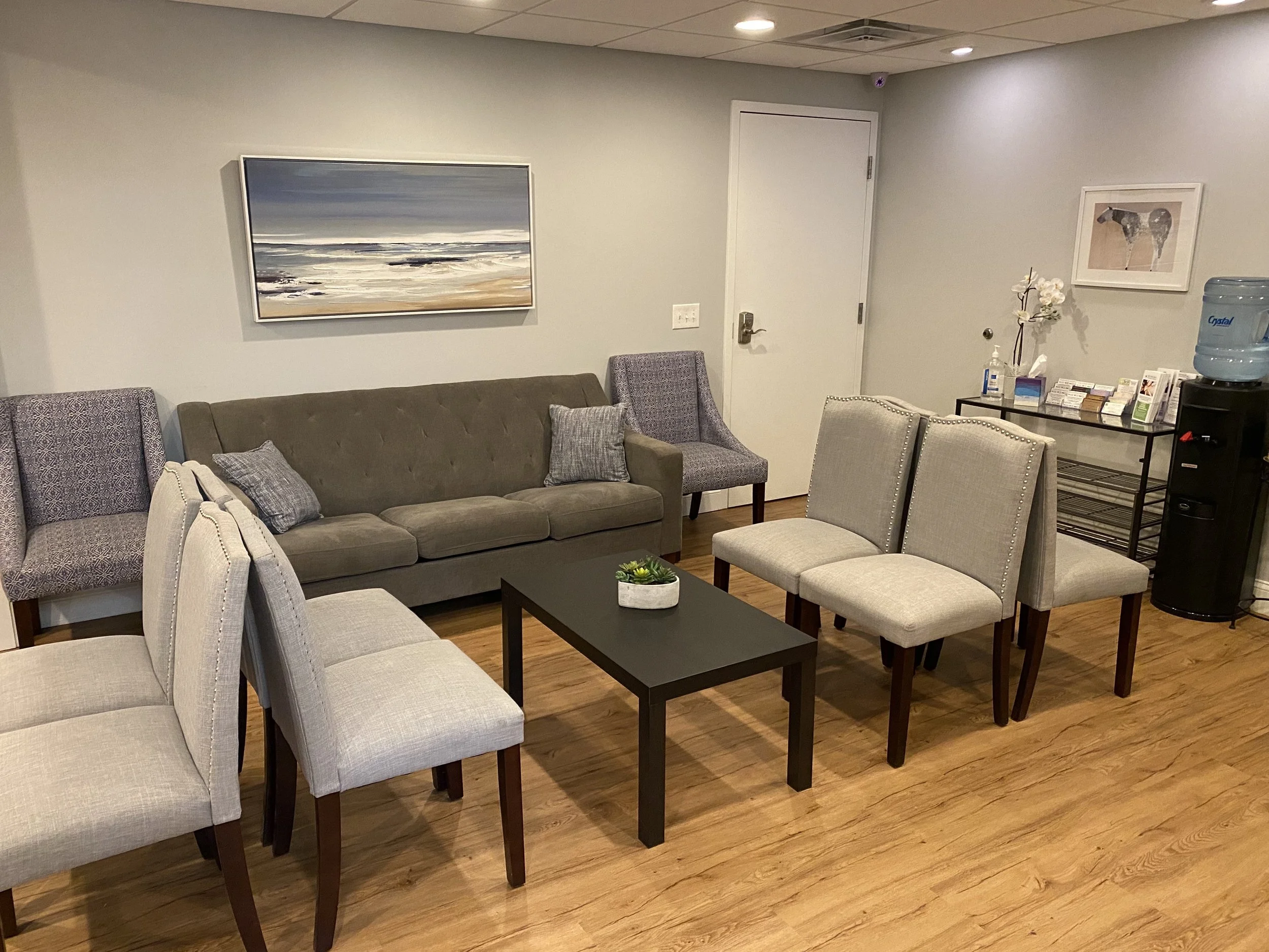 A waiting room with beige armchairs and a sofa, coffee table with a plant, framed artwork of a seascape on the wall, water cooler, tissue box, hand sanitizer, and brochures on a side table.