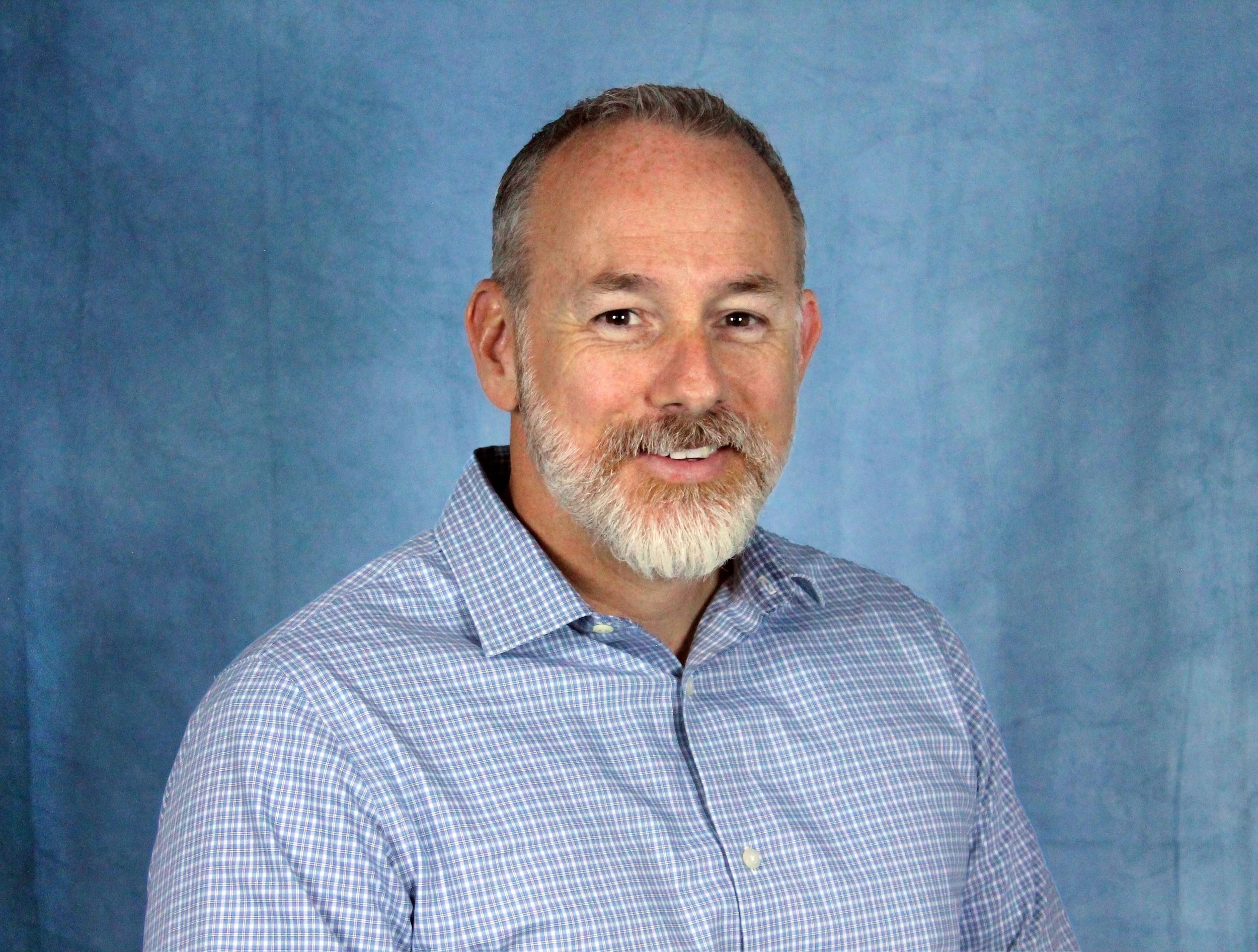 A middle-aged man with gray hair, a beard, and mustache wearing a blue checkered shirt, smiling against a plain blue background.