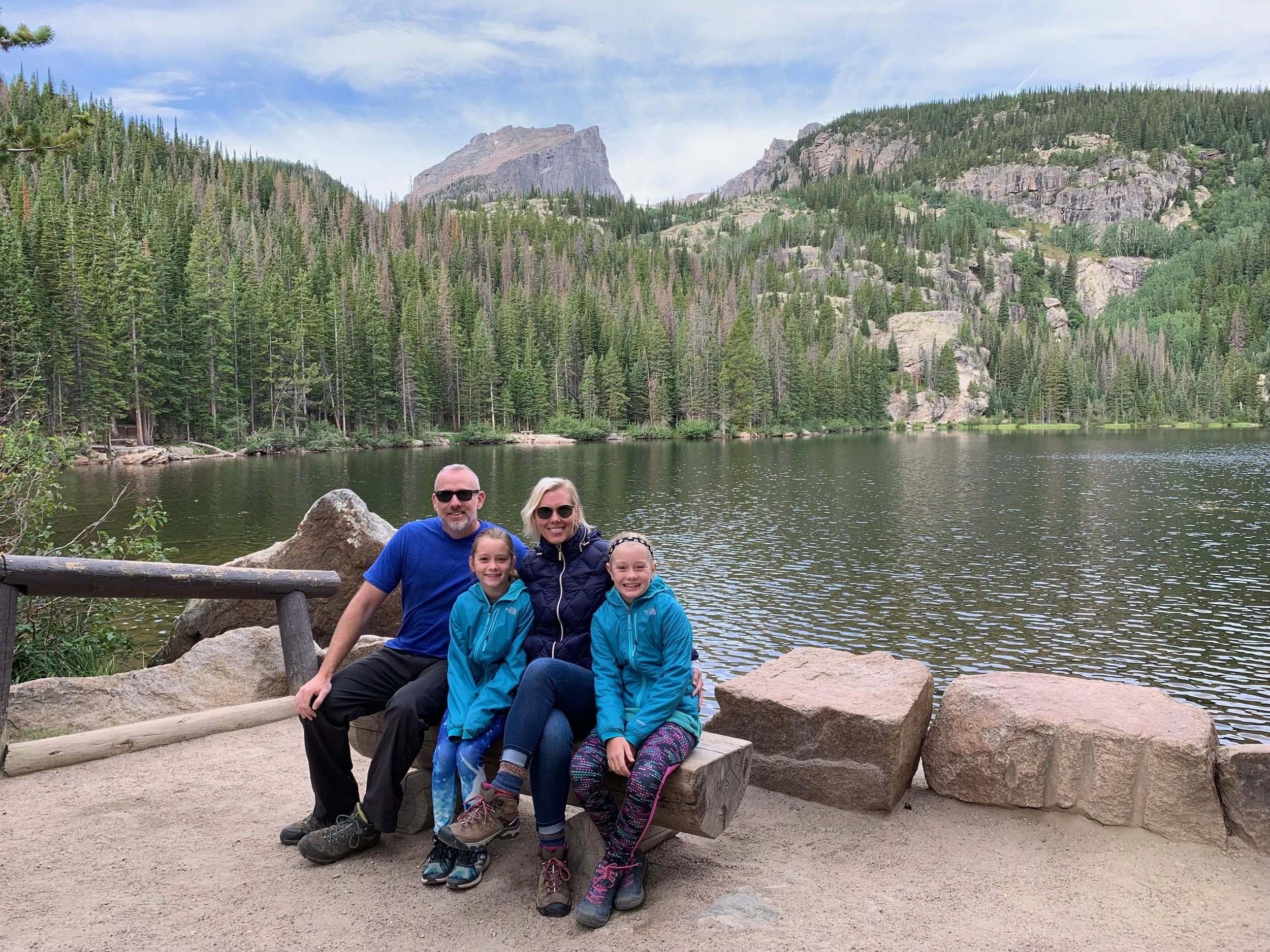 A group of five people, three girls and two adults, sitting on a large rock in front of a lake surrounded by dense pine forest and mountains under a cloudy sky.