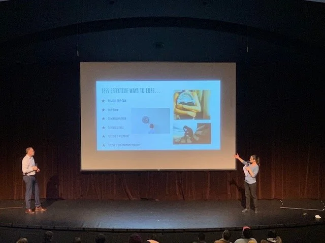 Two presenters on stage in front of an audience presenting a slide about managing stress with images of a hammock and a person relaxing on a beach.