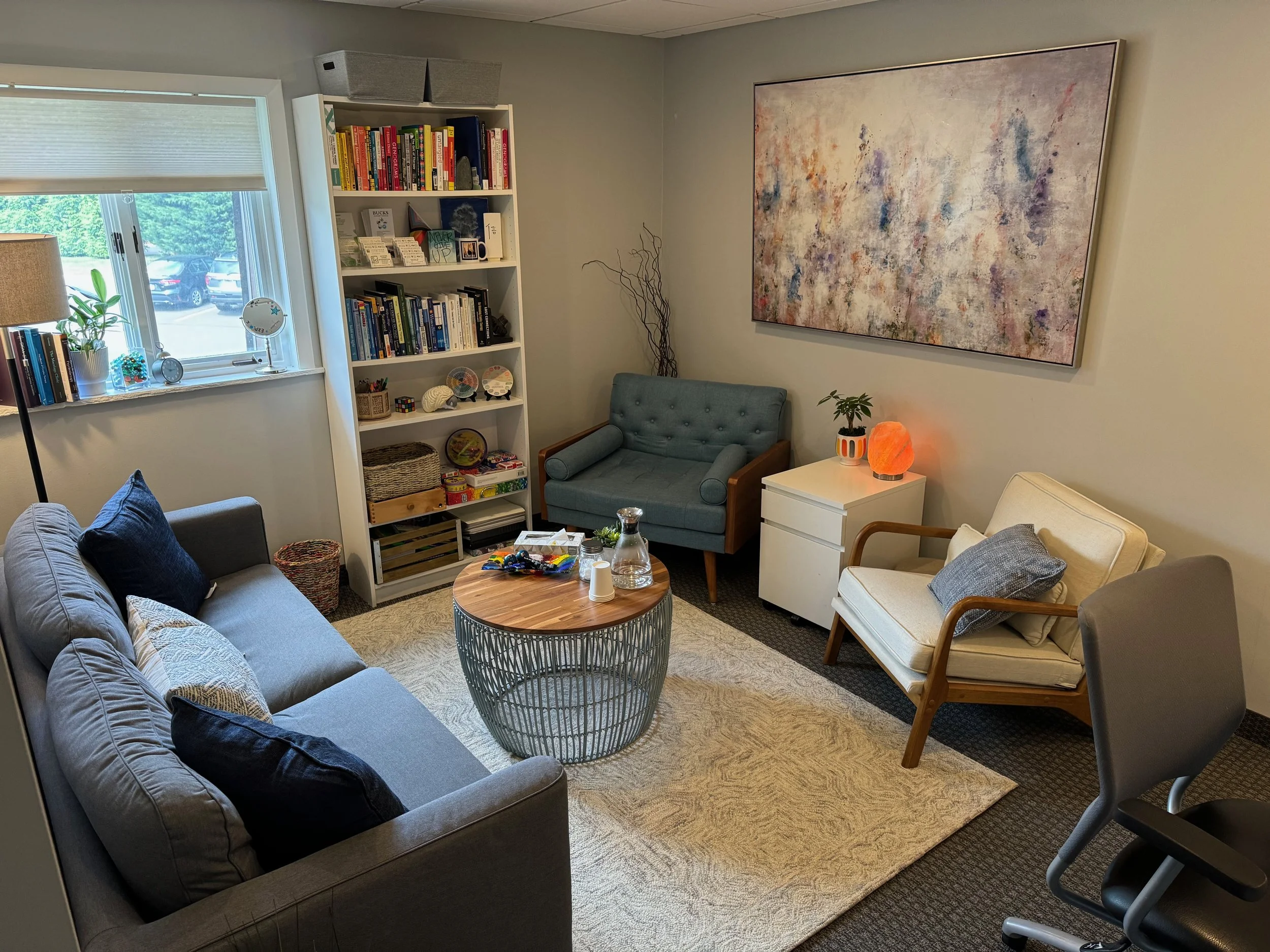 Living room with a gray sofa, a blue armchair, a white armchair, a round coffee table, a tall bookshelf filled with books and decorations, a window with blinds, and wall art, with various small decorative items on tables and shelves.