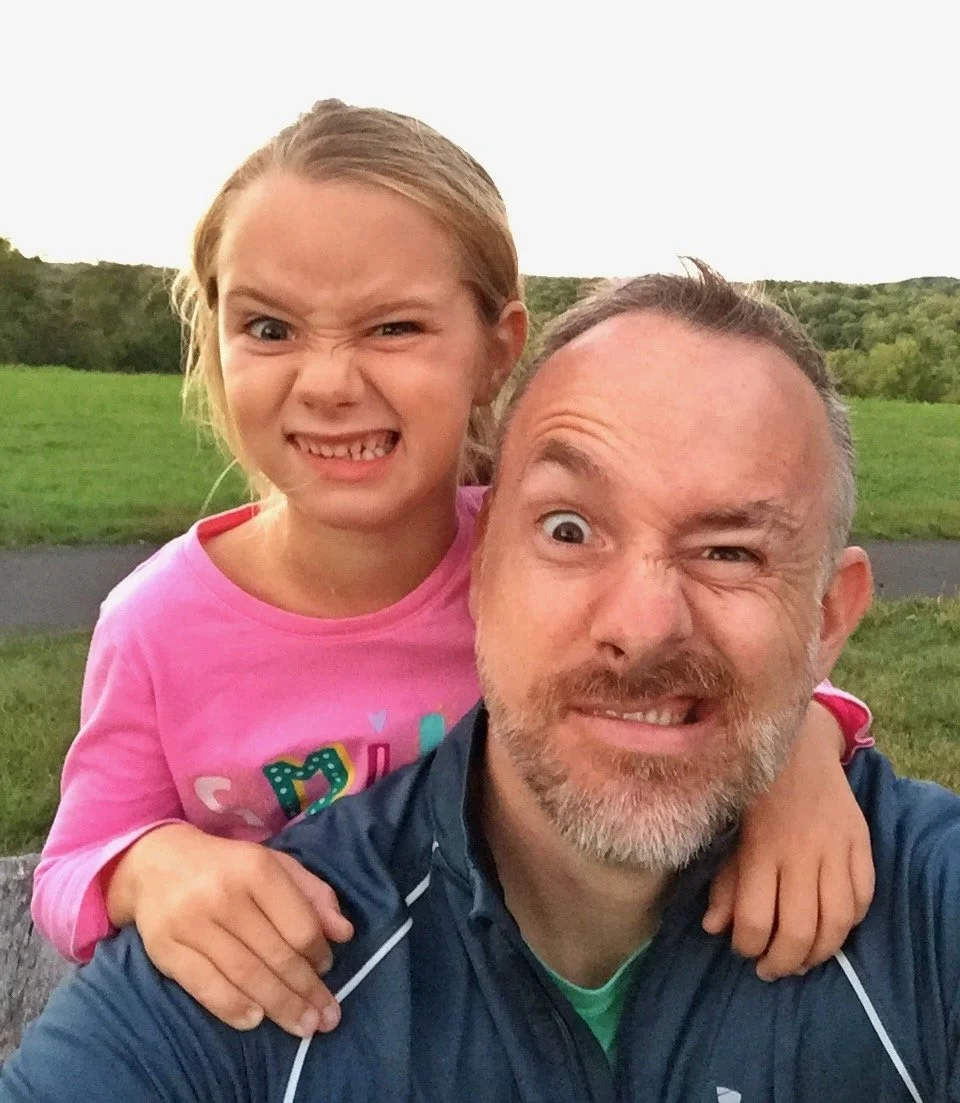 A man and a young girl are taking a selfie outdoors with a grassy field and trees in the background. The girl is wearing a pink shirt and making a funny face, while the man is smiling and squinting.