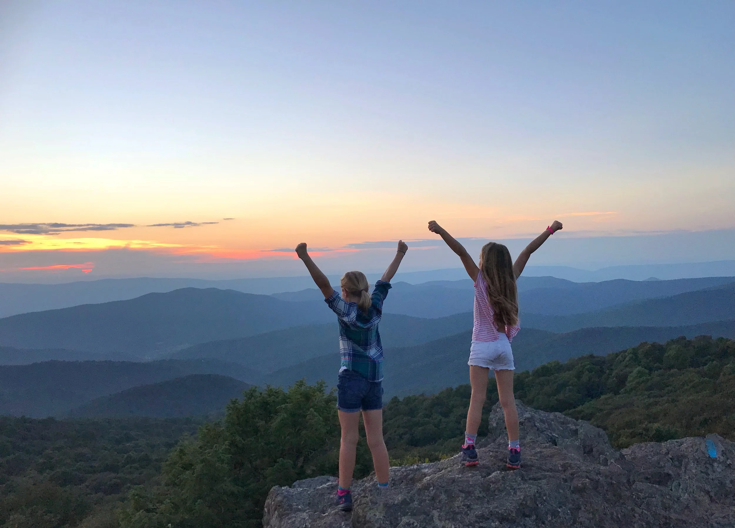 Two children are standing on a mountain ledge with their arms raised in celebration, overlooking a panoramic view of mountain ranges during sunset.