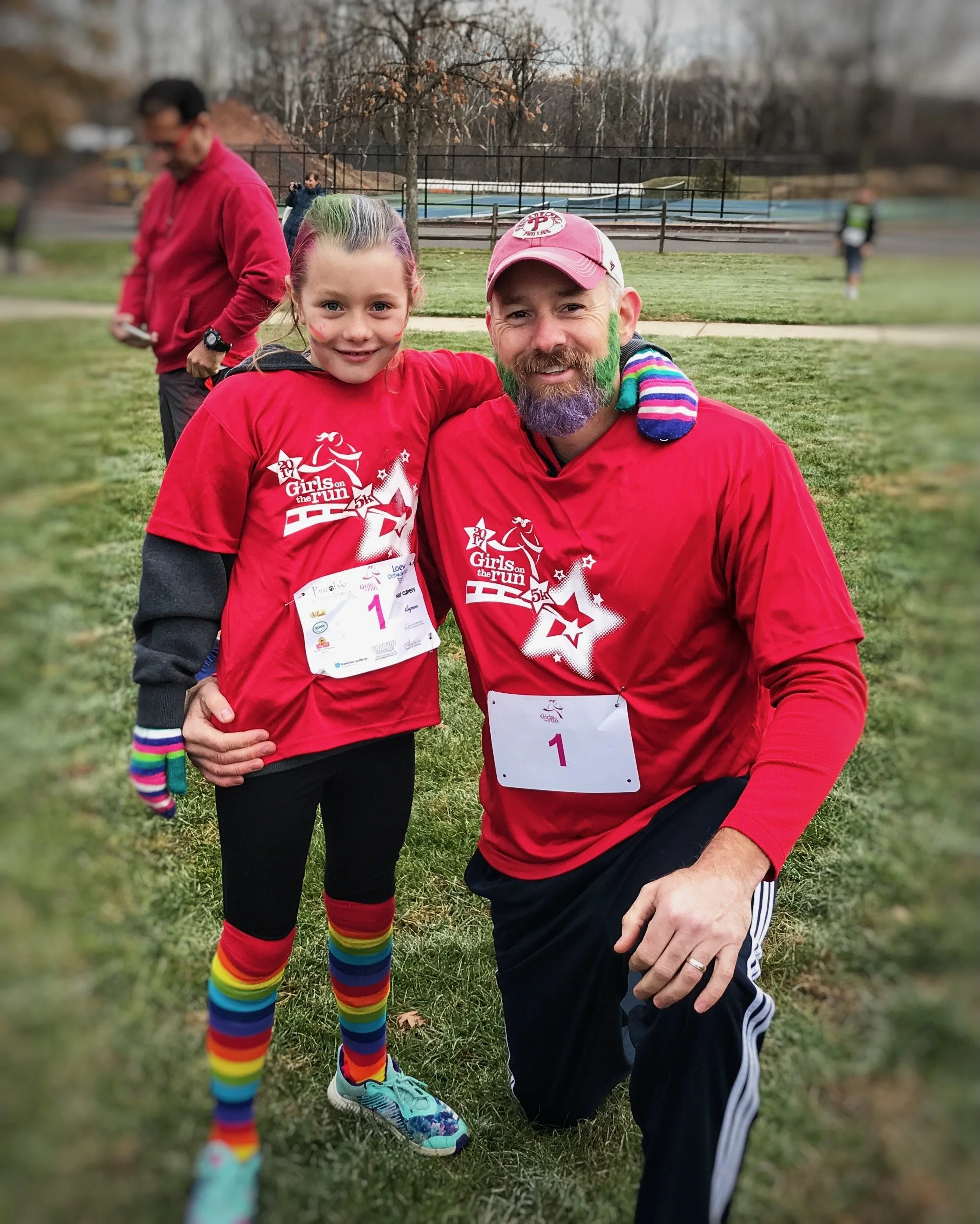 A young girl and an adult man, both smiling, taking a knee together at a park after a race event. The girl is wearing a red race shirt, rainbow-striped socks, and running shoes, with face paint. The man has a colorful beard, pink cap, and matching race shirt. In the background, there are other people and a tennis court, with autumn trees and overcast sky.