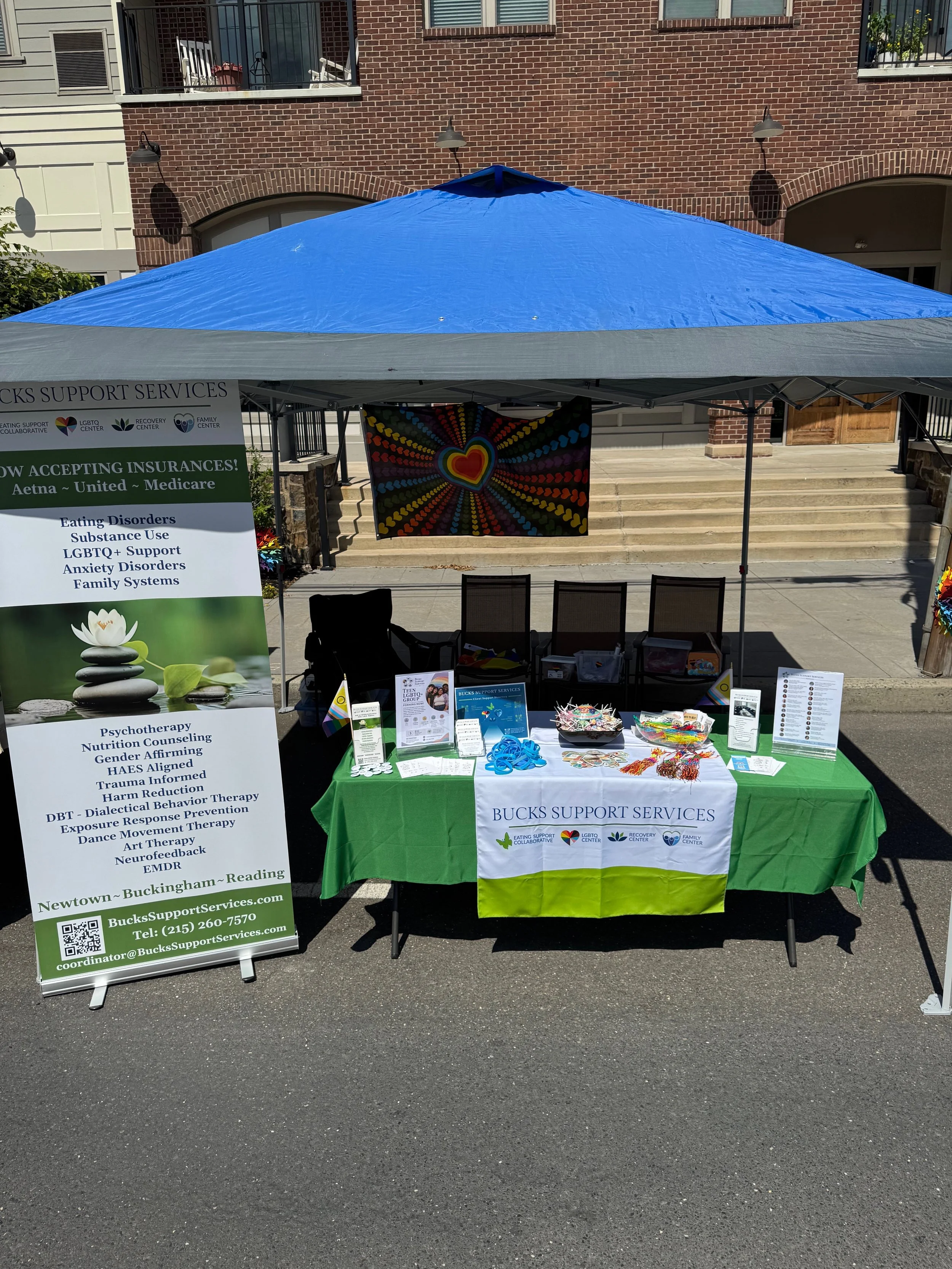 A support services booth set up outdoors with a blue canopy tent. The table is covered with a green and white tablecloth displaying the logo 'Bucks Support Services' and several informational pamphlets, decorative items, and rainbow-colored accessories. A large vertical banner advertises services including mental health support, LGBTQ+ support, counseling, and other mental health services. Behind the booth, there's a colorful rainbow heart tapestry hanging from the tent and a brick building facade with windows and balconies.