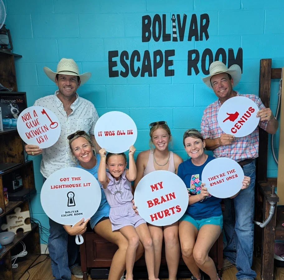 A group of seven people, four women and three men, are posing in front of a bright blue wall with a sign that reads 'Bolivar Escape Room.' They are holding various signs with phrases like 'Not a clue between us!!,' 'It was all me,' 'Genius,' 'Got the