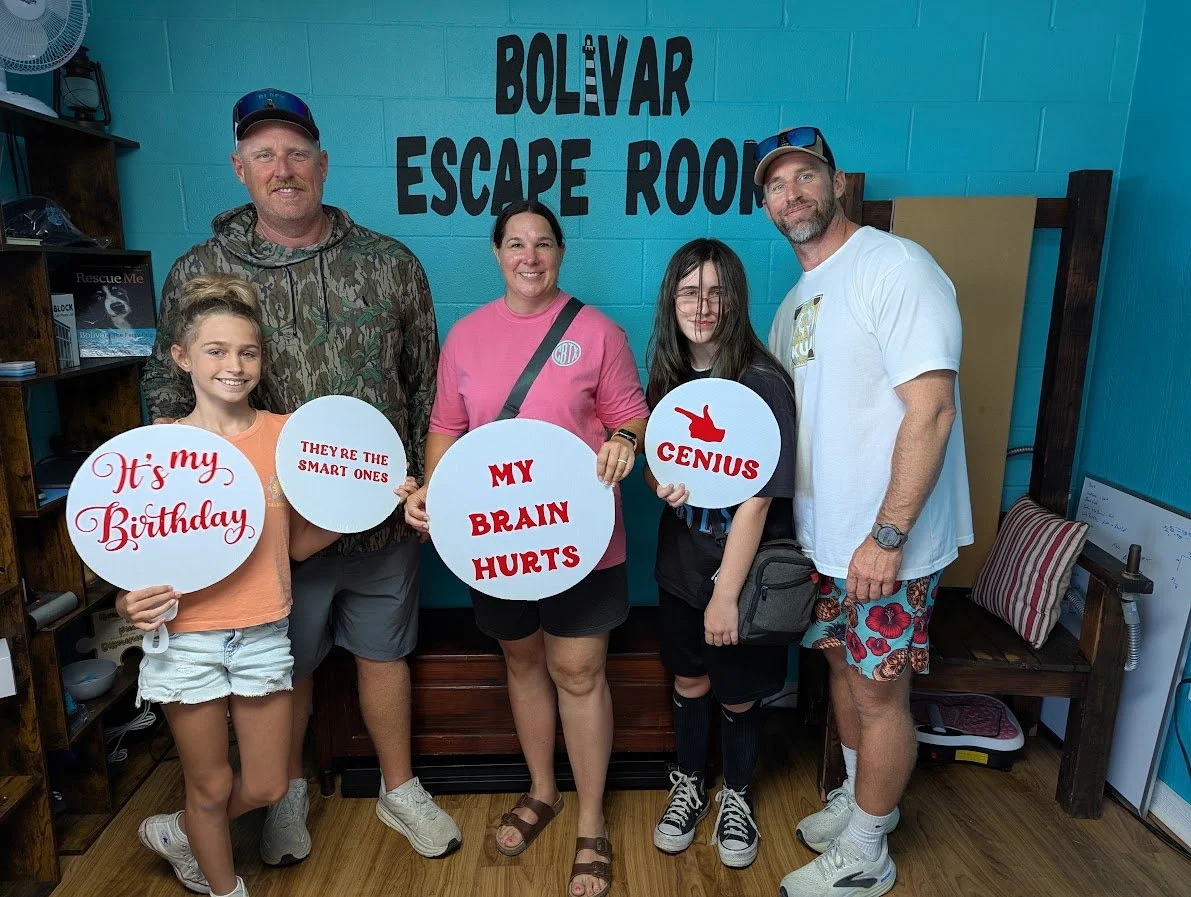 Group of five people standing inside an escape room called 'Bolivar Escape Room,' holding signs with humorous messages about brains and smartness, and one girl holding a sign that says 'It's my Birthday.'