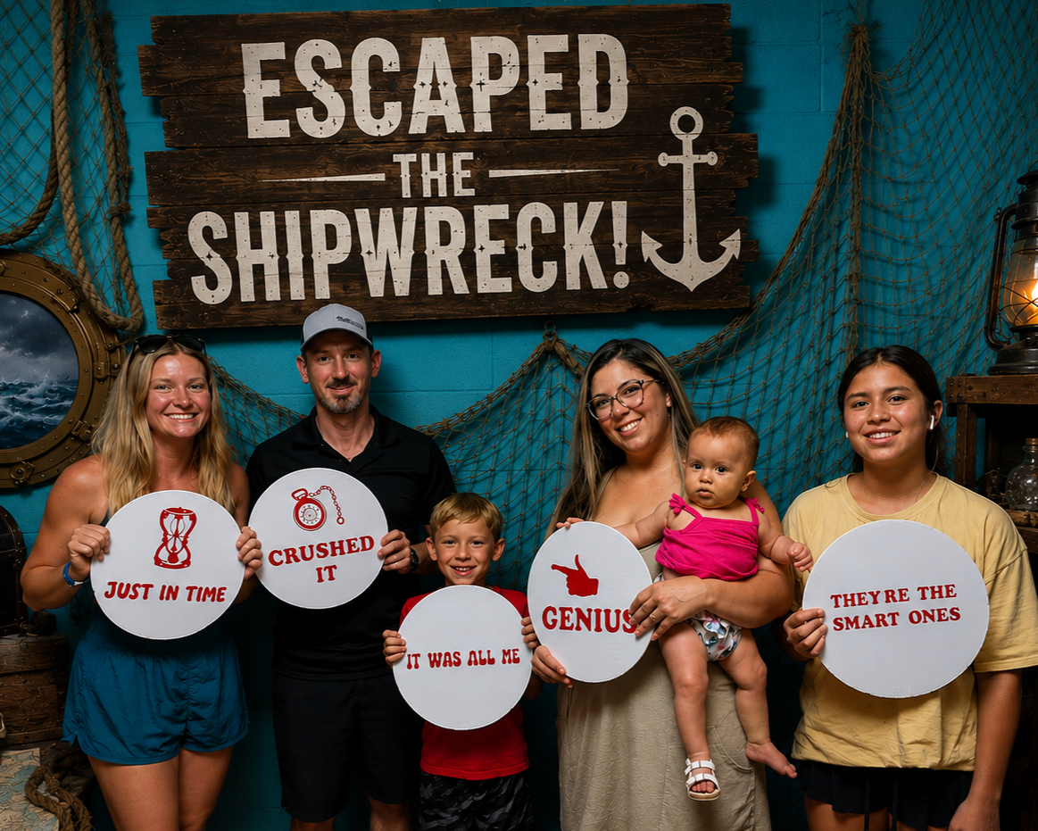 A group of six smiling people, including two children, pose in front of a nautical-themed backdrop with a sign that reads "Escaped the Shipwreck!" and hold signs with humorous messages, celebrating a fun event or party.