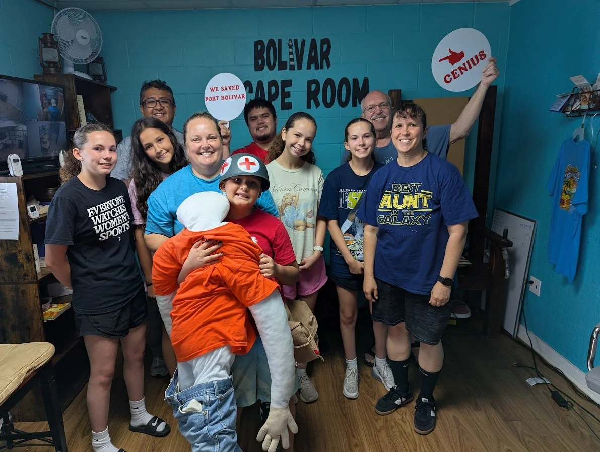 A group of people, including children and adults, are gathered inside a room at Bolivar Cape Room, with some holding signs. There is a person dressed in a costume resembling a chicken or a bird in the front. The background features a turquoise wall w