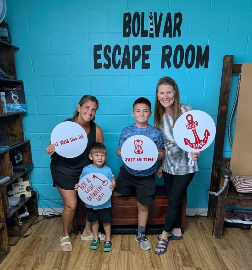 Four people, two women and two boys, holding signs, standing in front of a blue wall with a sign that reads 'Bolivar Escape Room'.